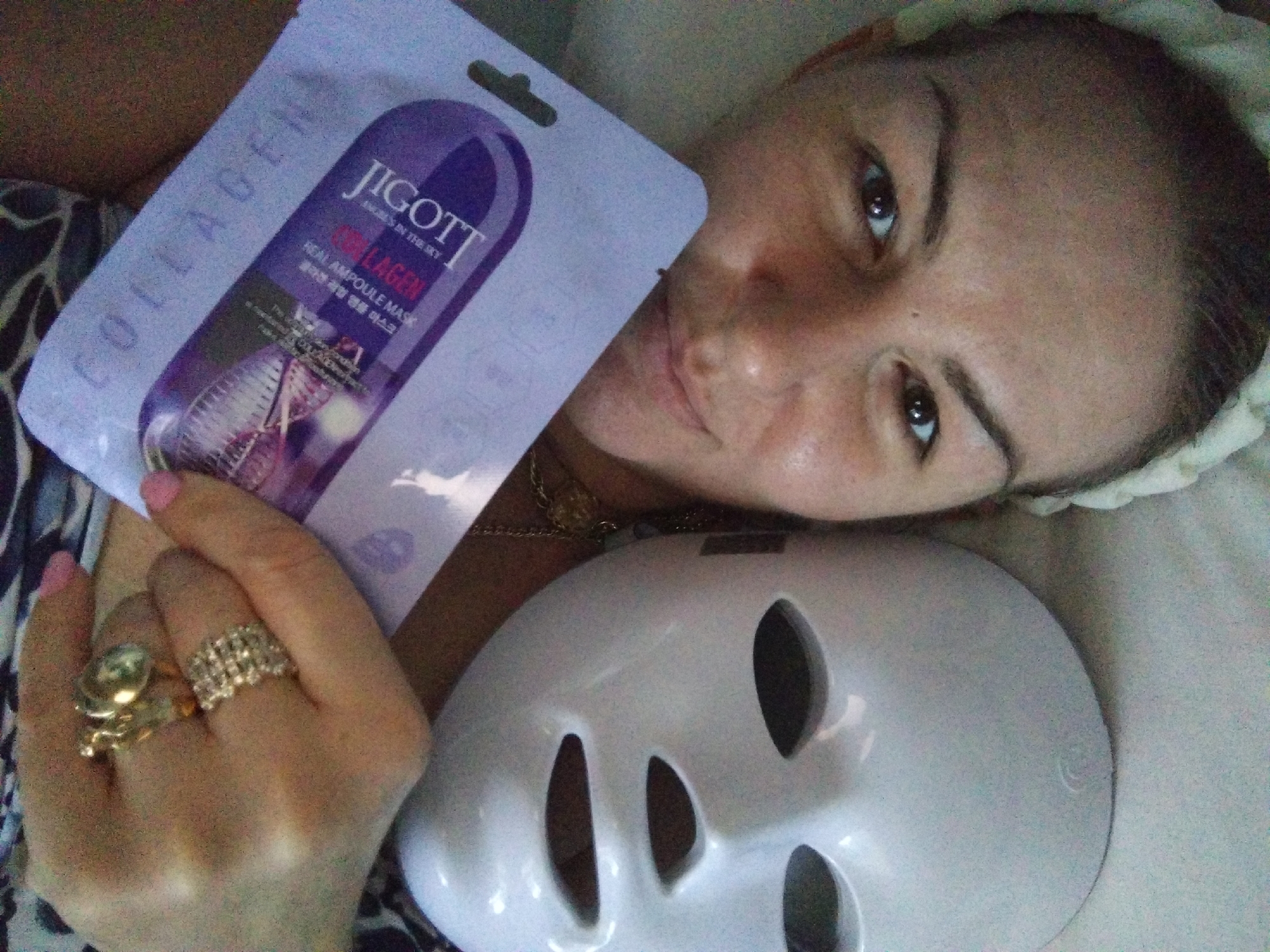 A close-up, overhead shot shows a woman lying down, her face tilted upwards. She is wearing a white LED facial mask with three eye openings and two larger openings for the nose and mouth. A sheet mask packet, labeled "Jigott REAL AMPOULE MASK COLLAGEN," is held over her chest, partially obscuring it. Her left hand, adorned with two rings, is visible near the sheet mask. The woman has dark hair, prominent eyebrows, and brown eyes. The background appears to be a light-colored, textured surface. The lighting is soft, suggesting an indoor setting. The image does not provide any information about the city of Stara Zagora, Bulgaria.