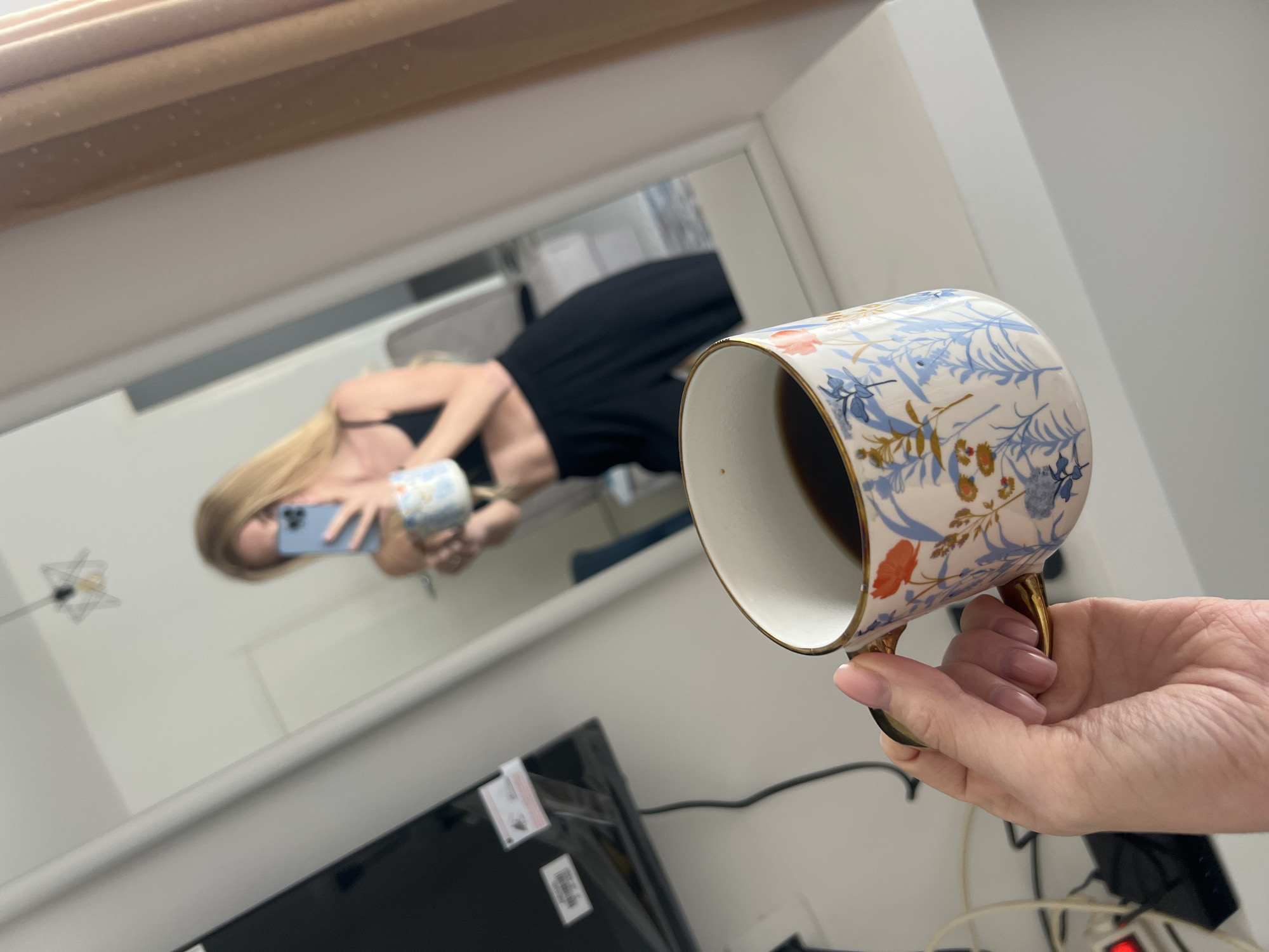 The image captures a mirror selfie taken indoors, likely in Budva, Montenegro, with a strong focus on a hand holding a coffee cup in the foreground. The perspective is tilted, with the top of the image angled upwards to the left.
In the foreground, a fair-skinned hand with long, light-colored fingernails holds a white ceramic coffee cup. The cup features an intricate floral pattern in shades of blue, orange, and gold, with a golden rim and handle, and contains a dark liquid, presumably coffee.
Reflected in the mirror, a blonde woman wearing a black top or dress is visible, holding a light blue smartphone in her left hand, which she uses to take the photo. Her right hand, also reflected, holds another patterned coffee cup, similar in style to the one in the foreground but appearing lighter in color. The reflection shows parts of a room with white walls and a light-colored wooden structure above the mirror, possibly a shelf or window frame. Below the mirror's reflection, the top portion of a black television or monitor is visible, displaying two white stickers with barcodes and unreadable text, one with a small red warning symbol. A tangled mess of cables and a power strip with a red light are also partially visible in the lower right background, behind the hand holding the cup. The overall scene suggests a casual, indoor moment, possibly morning given the presence of coffee.