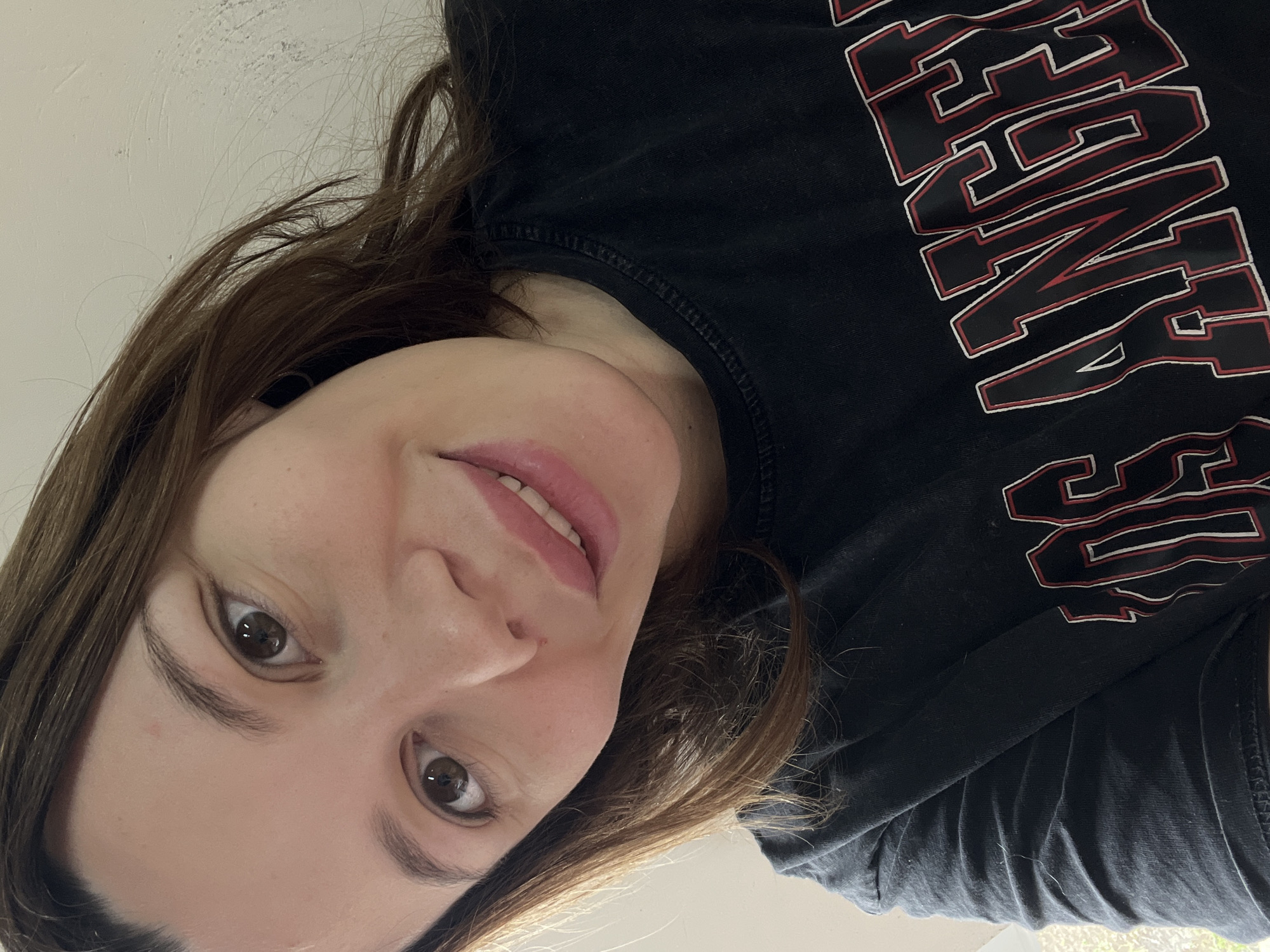 A close-up, slightly upward-angled indoor portrait depicts an adult female with fair skin, medium-length brown hair, and brown eyes. She wears a black short-sleeved t-shirt featuring a partially visible "LOS ANGELES" graphic in white outline with red interior lettering. A faint silver-colored necklace chain is visible around her neck. The subject has a slight smile, showing a portion of her upper teeth, and gazes directly into the camera. The background is a plain light-colored wall, with an indistinct light source or window situated to the subject's left. No discernible features indicate the city of Leninakan, Russia, or any specific geographic location.