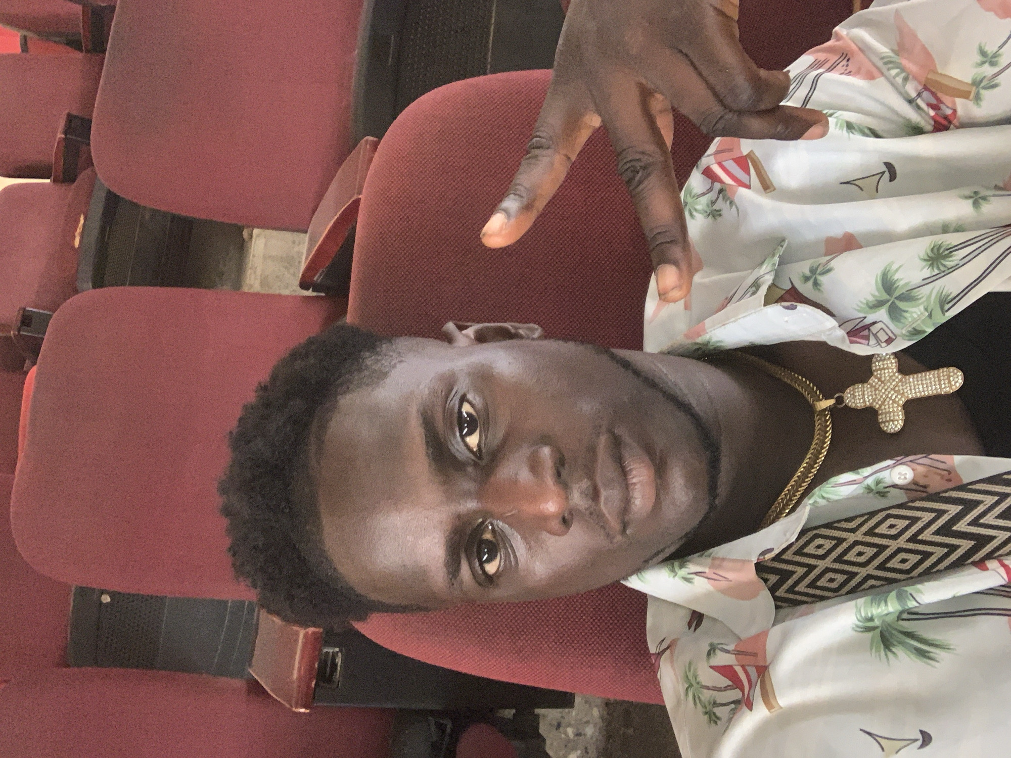 A young Black man with short, dark, curly hair and dark skin is seated in a red theater seat, looking towards the camera. He is wearing a white collared shirt with a tropical print featuring palm trees and sailboats. He has a thick gold chain around his neck with a large, diamond-encrusted cross pendant. His right hand is raised, with his index and middle fingers extended in a peace sign. The background consists of more red theater seats, suggesting an indoor venue. The lighting is consistent with an interior setting, possibly a movie theater or auditorium, and there are some visible dust or debris on the floor between the seats. The man appears to be indoors in Jalingo, Nigeria.