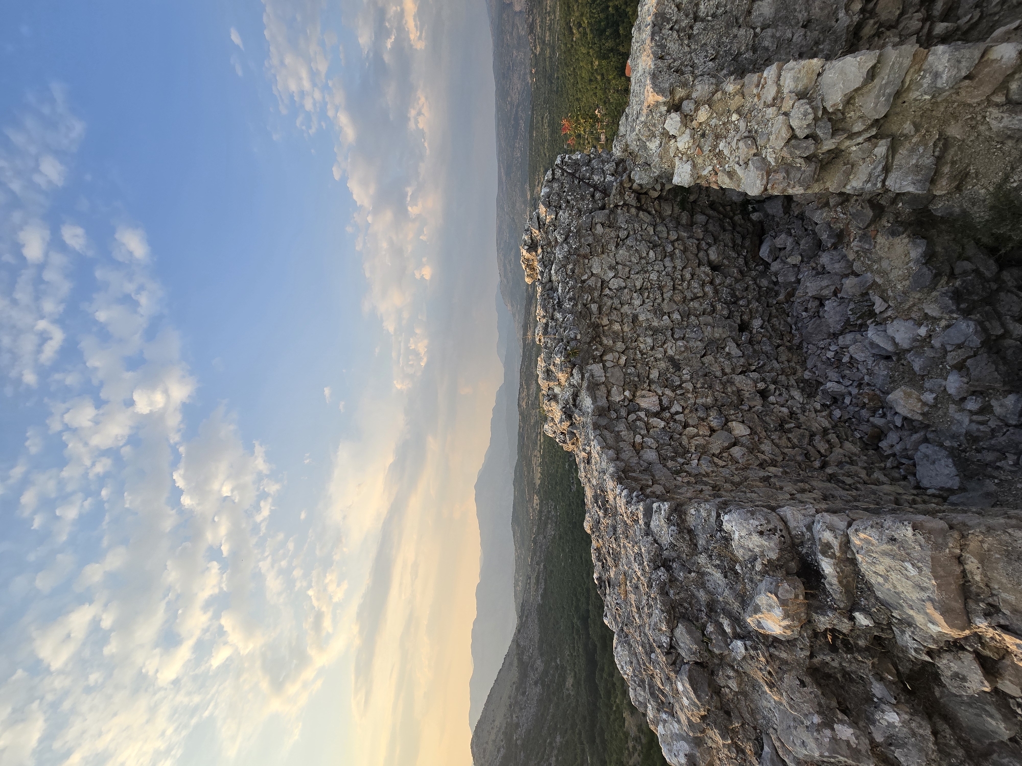 The image depicts a stone ruin structure overlooking a valley with mountains in the distance. The sky above is blue with scattered white clouds, illuminated by what appears to be late afternoon sunlight. The ruin is made of rough-hewn stones, some of which are loose and have fallen into the interior of the structure. The surrounding landscape is covered in dense green vegetation, with some scattered trees and shrubs. The mountains in the background are hazy and appear to be layered, suggesting a vast expanse of land. This scene is likely captured from a vantage point within or near the old fortress of Medun, Montenegro, a historical site known for its ruins and panoramic views of the surrounding landscape. There are no people visible in the image.