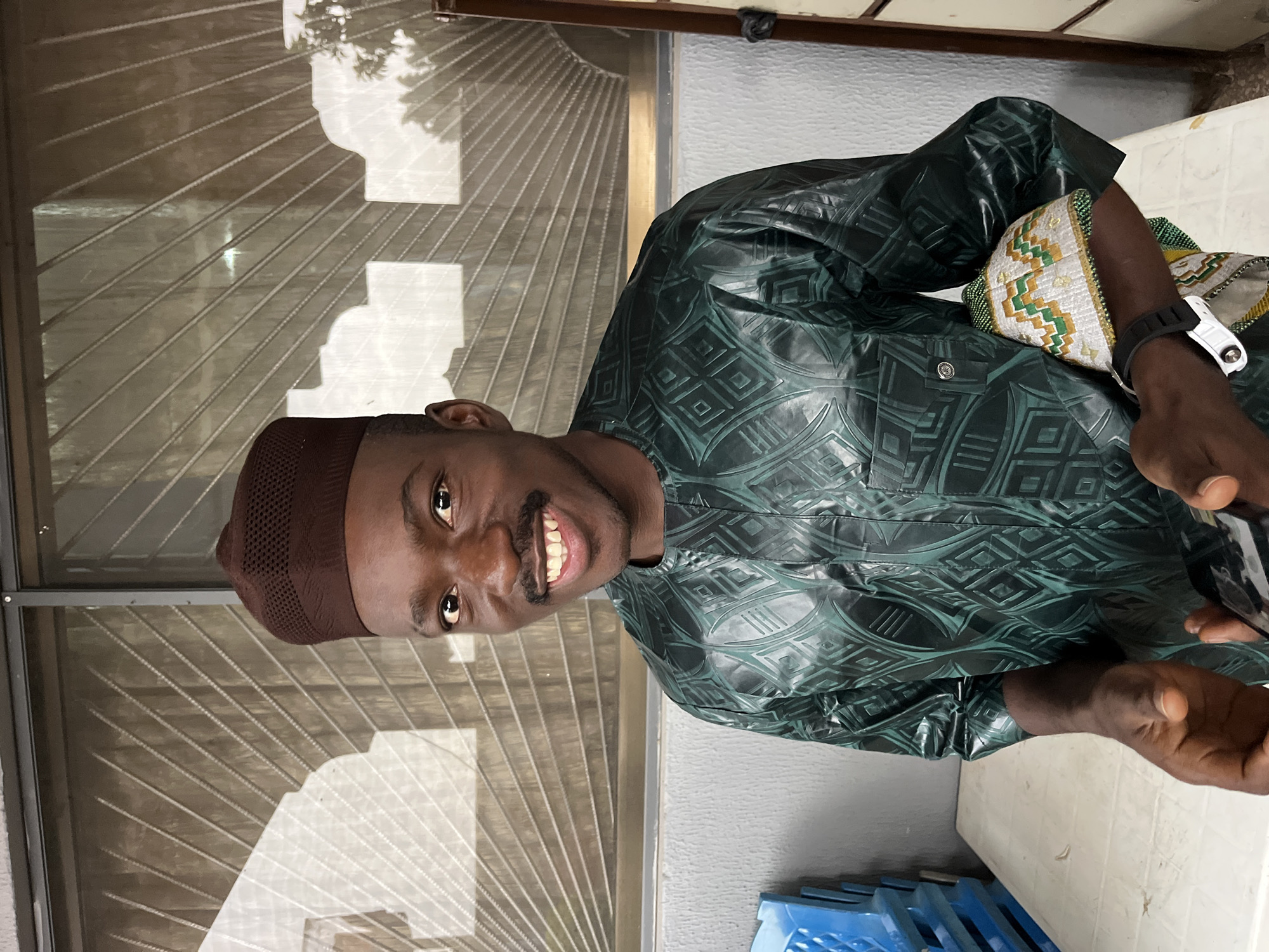 A young Black male, wearing a brown kufi and a dark green patterned traditional African garment, is pictured from the waist up, smiling directly at the camera. He has a mustache and is wearing a black watch on his left wrist. He is holding a phone and a patterned cloth in his hands. The background consists of a textured window with horizontal bars and reflections of outdoor foliage. To the left, blue plastic chairs are stacked, and a white table is in the foreground. The scene appears to be outdoors or in a covered outdoor area.