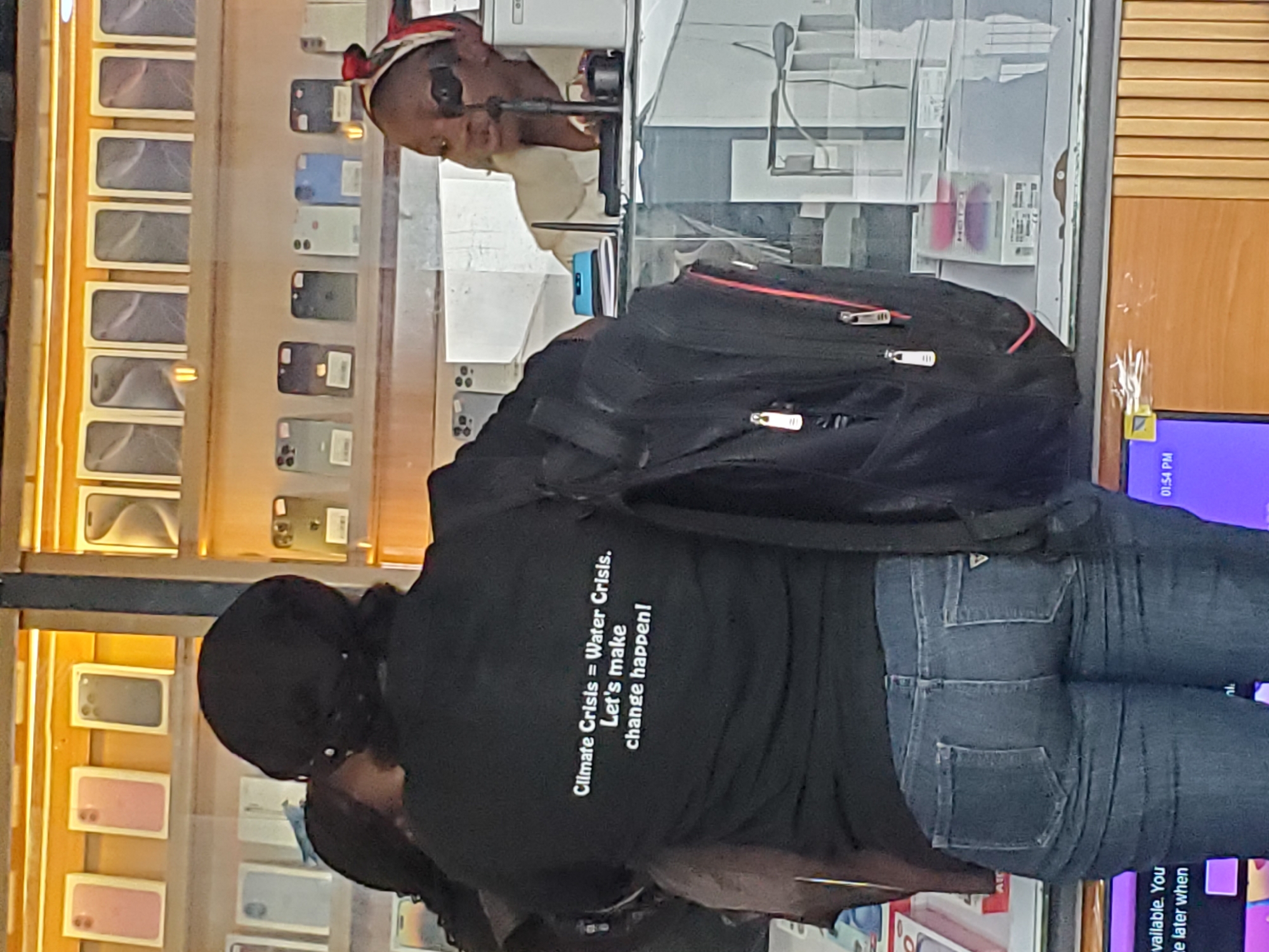 The image shows a person wearing a black t-shirt with white text on the back that reads "Climate Crisis = Water Crisis. Let's make change happen!". The person is also wearing blue jeans and has a black backpack. The scene appears to be an electronics store, with shelves filled with mobile phones and their boxes visible in the background. A screen displaying the time "01:54 PM" is also visible on the right side of the image, suggesting it is afternoon. The overall setting suggests a retail environment focused on consumer electronics. The person's attire and the message on their t-shirt imply a statement about environmental awareness. The context provided is Jos, Nigeria.