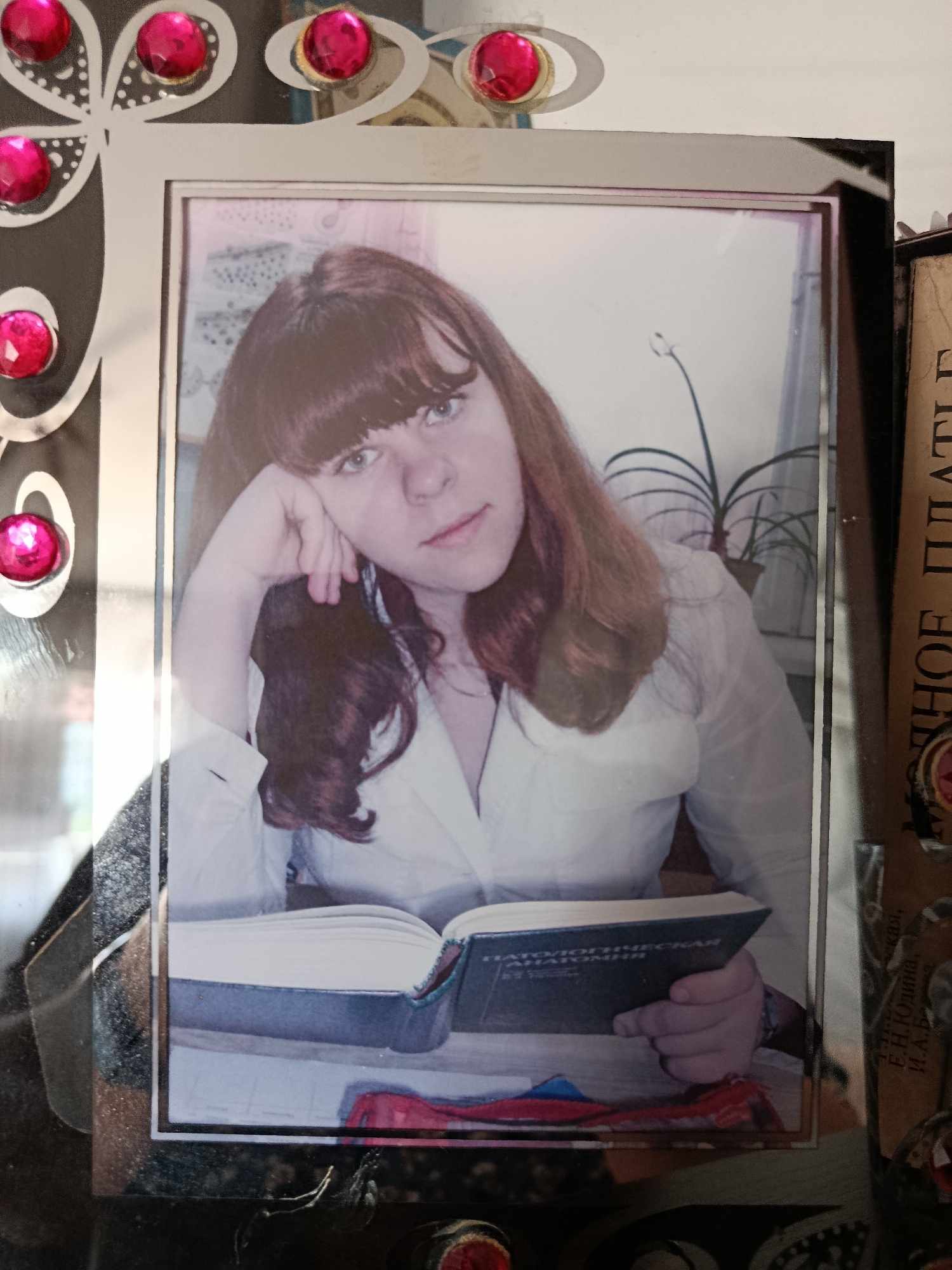 A candid, eye-level, slightly high-angle medium shot captures a young woman with auburn hair and bangs, leaning on her hand, in a photographic frame with decorative pink gems. She is wearing a light-colored button-down shirt and is looking towards the viewer. She is seated at a table with an open book in front of her, which she is holding. The background appears to be an indoor setting with a plant visible. The frame is positioned within a larger frame or display unit, which also shows part of a book with Russian text. The overall impression is a personal keepsake. The location is identified as Svietlahorsk, Belarus.