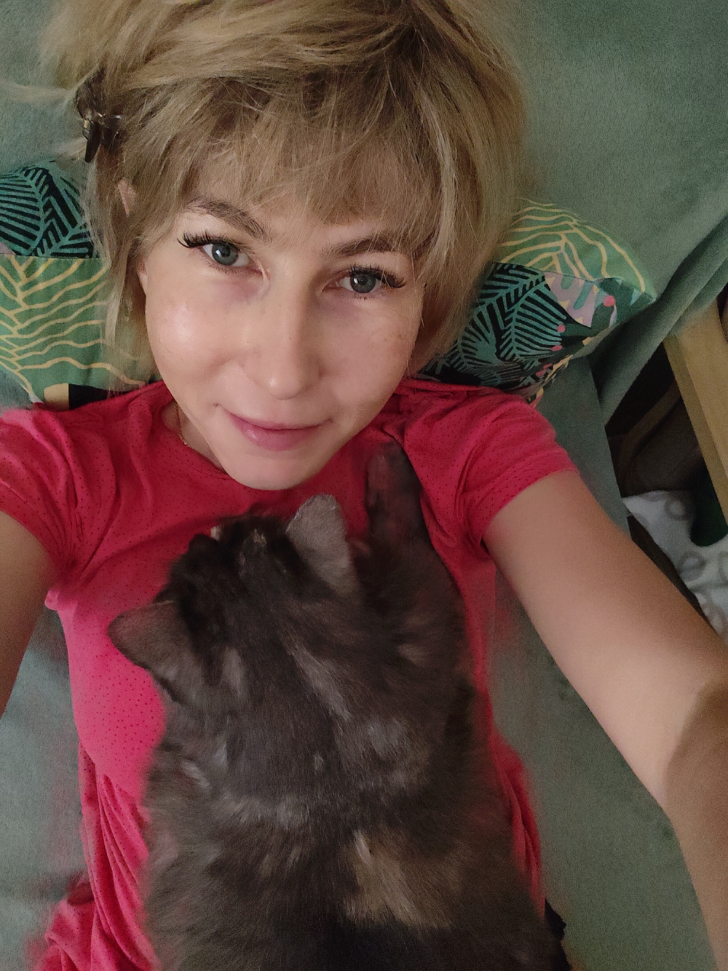 The media file is a selfie taken from above, featuring a young woman with blonde hair and blue eyes, lying down with a cat on her chest. She is wearing a bright pink t-shirt and has makeup on, including false eyelashes. The cat is a dark grey or black tabby, with its head turned away from the camera. The woman is smiling gently, looking relaxed and happy. She is lying on a teal-colored surface, possibly a sofa or bed, with a green pillow featuring a pattern of tropical leaves behind her head. The background is slightly out of focus, but appears to be an indoor setting. The lighting suggests it could be daytime. The overall mood is cozy and affectionate, highlighting the bond between the woman and her pet.