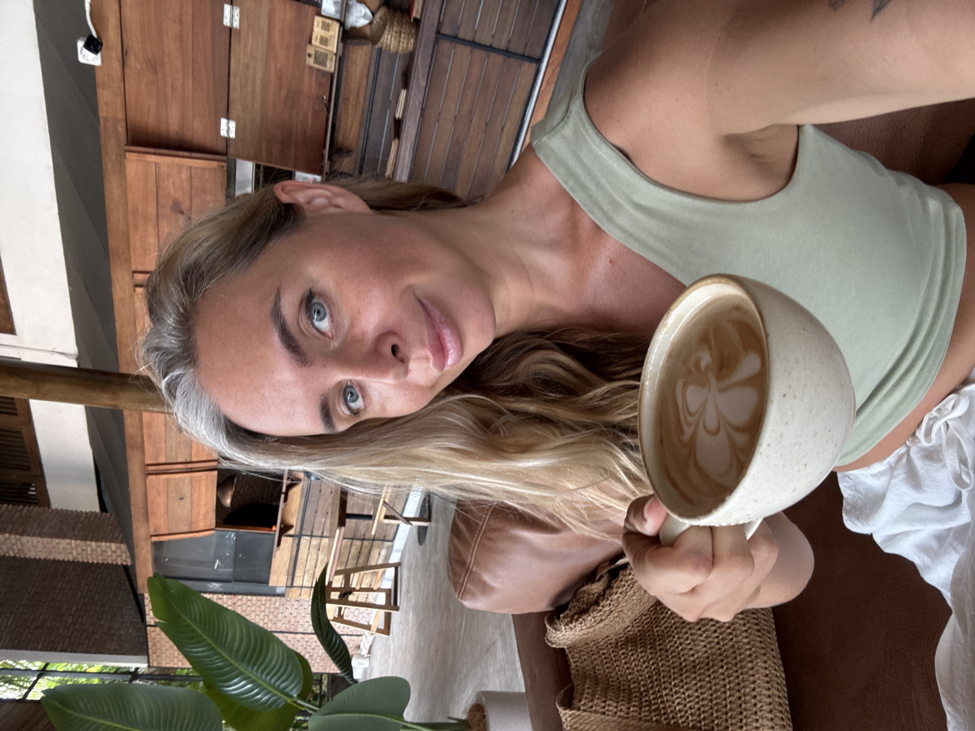 A woman with blonde hair and blue eyes is holding a cup of coffee with latte art in a cafe. She is wearing a light green tank top and white pants. The cafe has a rustic design with wooden furniture and brick walls. There are plants in the background, and a woven bag is placed on a brown leather sofa. The woman is looking at the camera and smiling. The scene appears to be in a cafe in Ko Samui District, Thailand.
