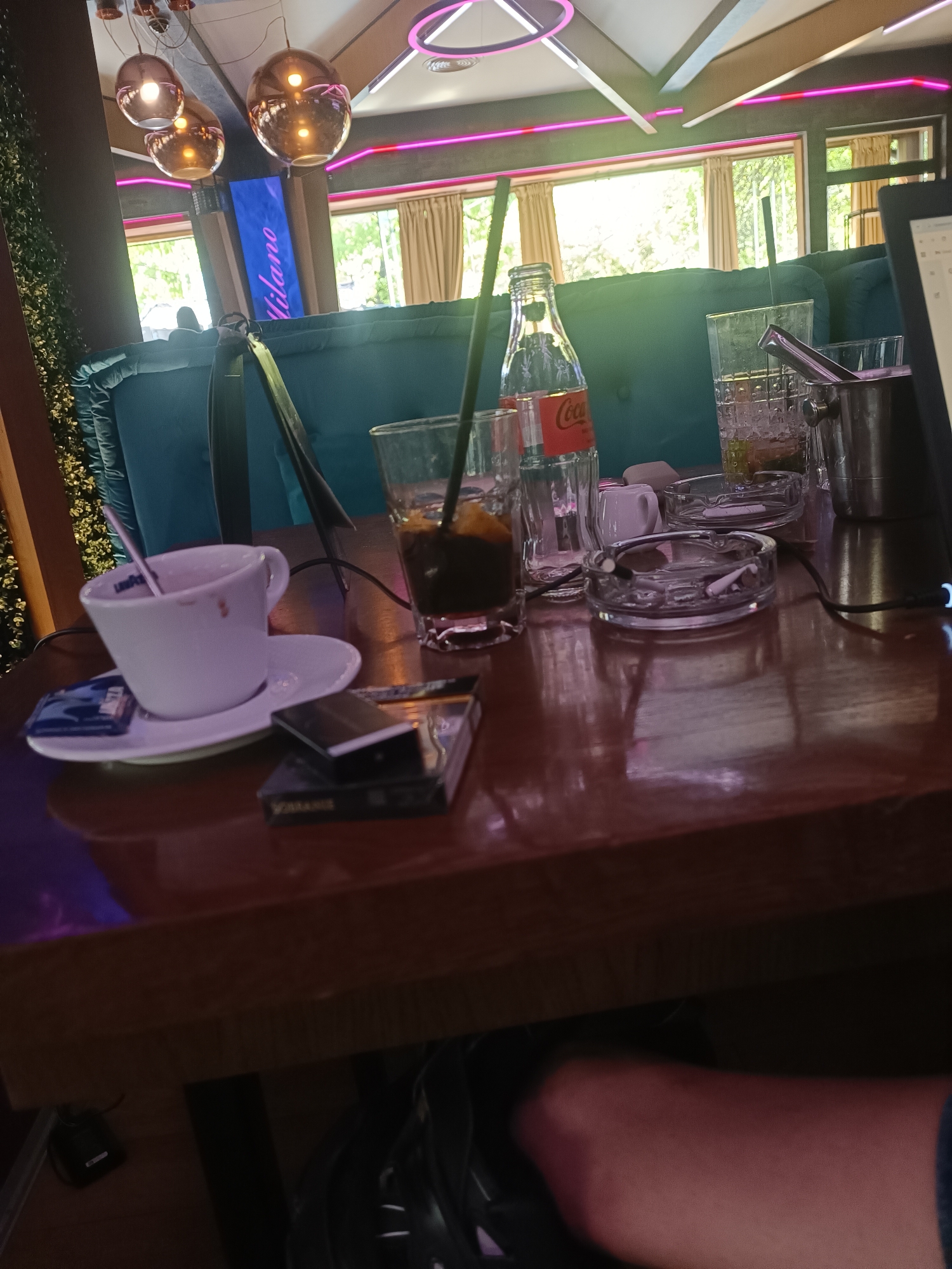 The image depicts a close-up of a table in what appears to be a cafe or bar. The table is cluttered with items including a half-empty cup of coffee on a saucer, a packet of sugar, a pack of cigarettes, a glass with a dark liquid and a straw, a glass Coca-Cola bottle, two ashtrays (one containing cigarette butts), and an ice bucket with tongs. Part of a person's leg is visible in the foreground. Outside the window, greenery and buildings are visible. A sign "Milano" is seen. The general decor suggests a relatively modern, upscale establishment. It is possible that the photo was taken in Sofia, Bulgaria based on the signage "Milano," which may imply a connection to Italy.