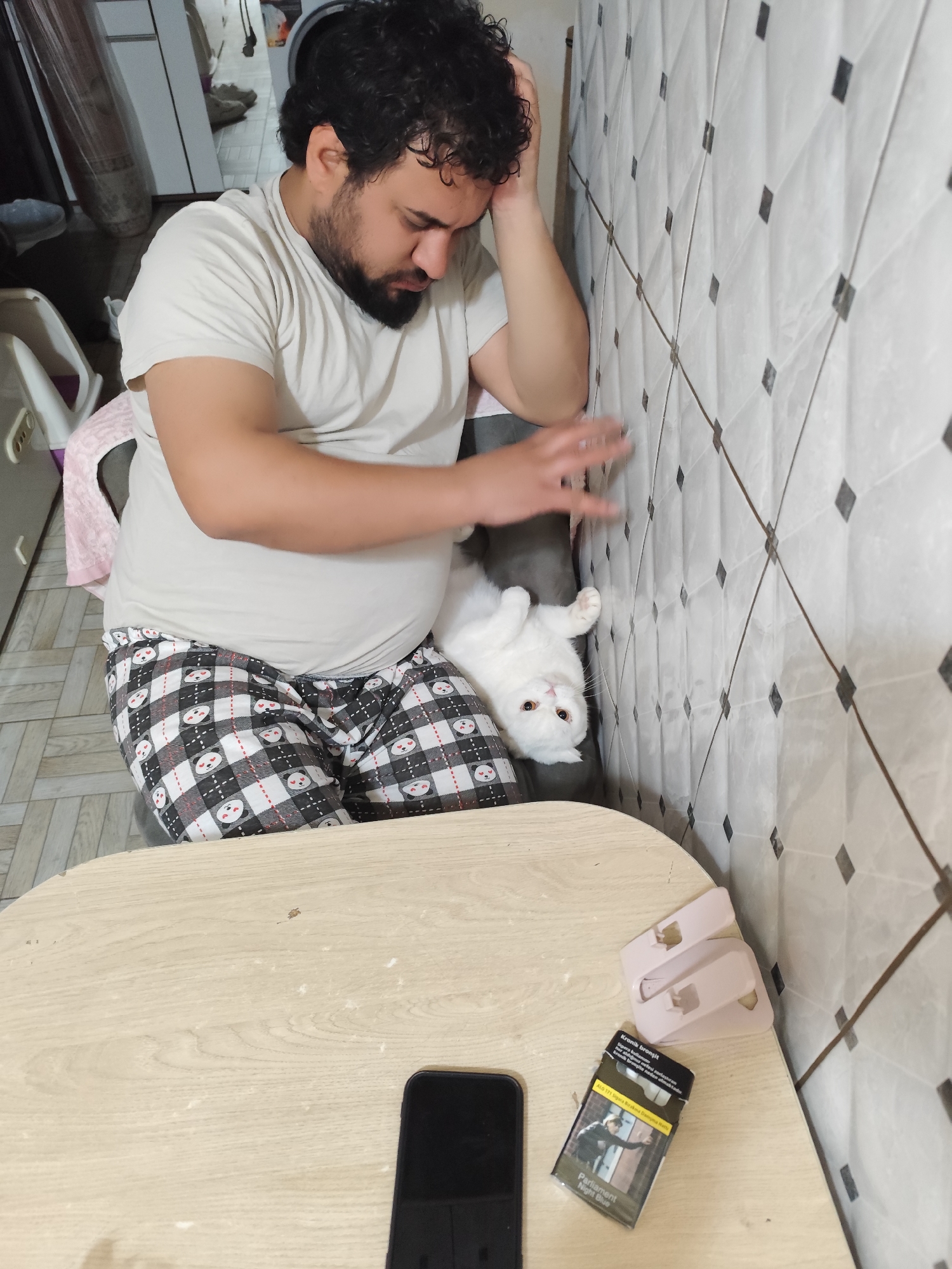 A man with dark curly hair and a beard sits in a chair with a white cat perched on his lap. The man wears a light tan t-shirt and pajama pants with a panda bear pattern. His right hand is raised and slightly blurred, suggesting movement. The cat is white with yellow eyes and looks directly at the camera. The background consists of a tiled wall. In the foreground, a light-colored wooden table holds a black smartphone, a pack of Parliament Night Blue cigarettes, and two light pink hair clips. The scene is indoors, possibly in a residential area in Sarıyer, Turkey.