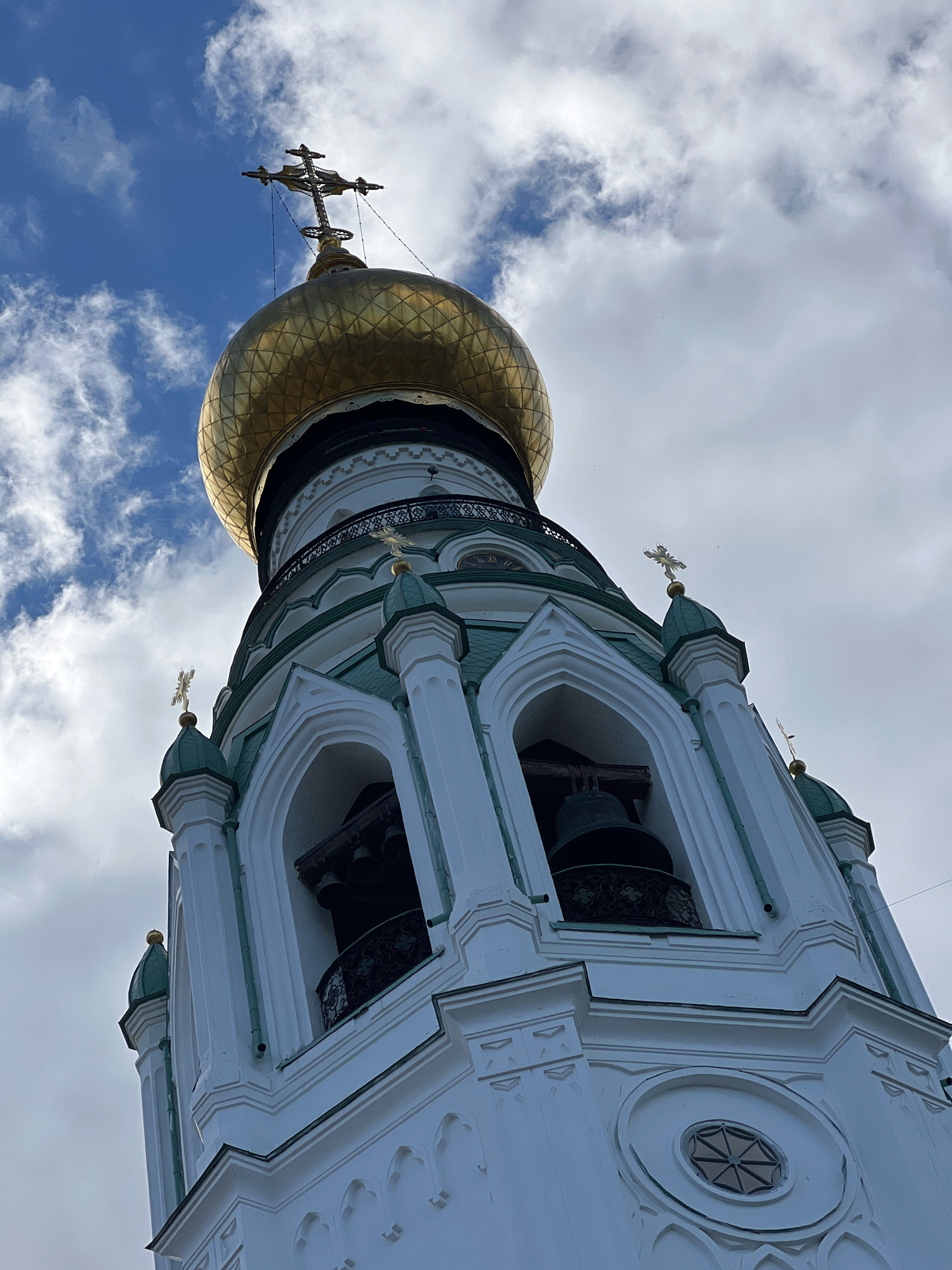 A low-angle shot shows the white bell tower of an Orthodox church against a cloudy blue sky. The tower has green accents and several arched openings where bells are visible. A golden dome with a cross tops the structure. The image is taken from the ground looking upwards, with the sky filling much of the frame.