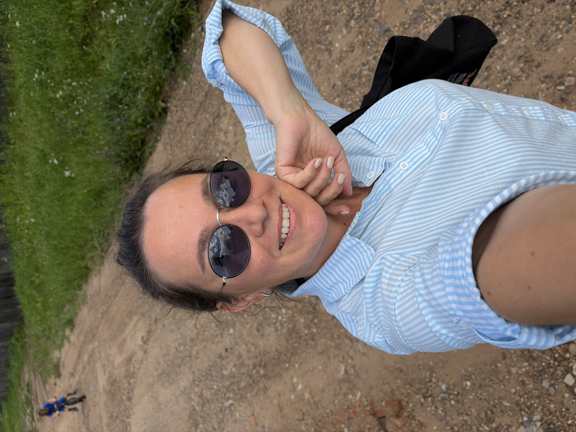 In a sun-drenched, outdoor setting, a woman with dark hair pulled back smiles at the camera, her hand resting on her cheek. She wears a light blue and white striped button-down shirt and large, round sunglasses. To the left and in the background, a young child in a blue shirt rides a bicycle on a dirt path lined with dry grass and occasional green vegetation. The ground is a mix of dirt and small stones. A wooden fence is visible in the far distance, bordering a field of tall green grass. The scene appears to be in a rural or semi-rural area, possibly the city of Olkhovka, Russia, given the natural landscape and the simple wooden fence.