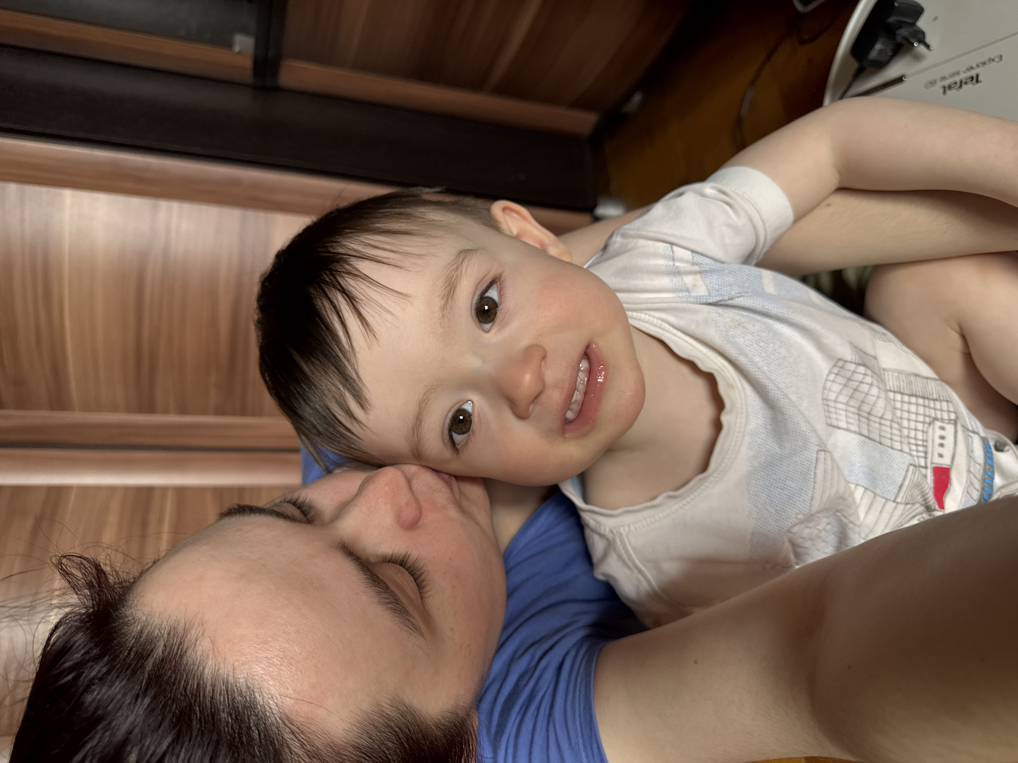 A close-up, angled shot depicts an adult woman lying down with a toddler resting on her chest. The woman has dark hair pulled back and is wearing a blue t-shirt. Her eyes are closed, and she appears to be kissing the child's head. The toddler, with dark hair and brown eyes, is looking directly at the camera. They are wearing a white t-shirt with a print of what appears to be buildings. The background includes a wooden-paneled wall and an out-of-focus object that reads "Tefal" in white letters on the upper right corner. The location in the city of Izhevsk, Russia cannot be determined from the details provided in the image.