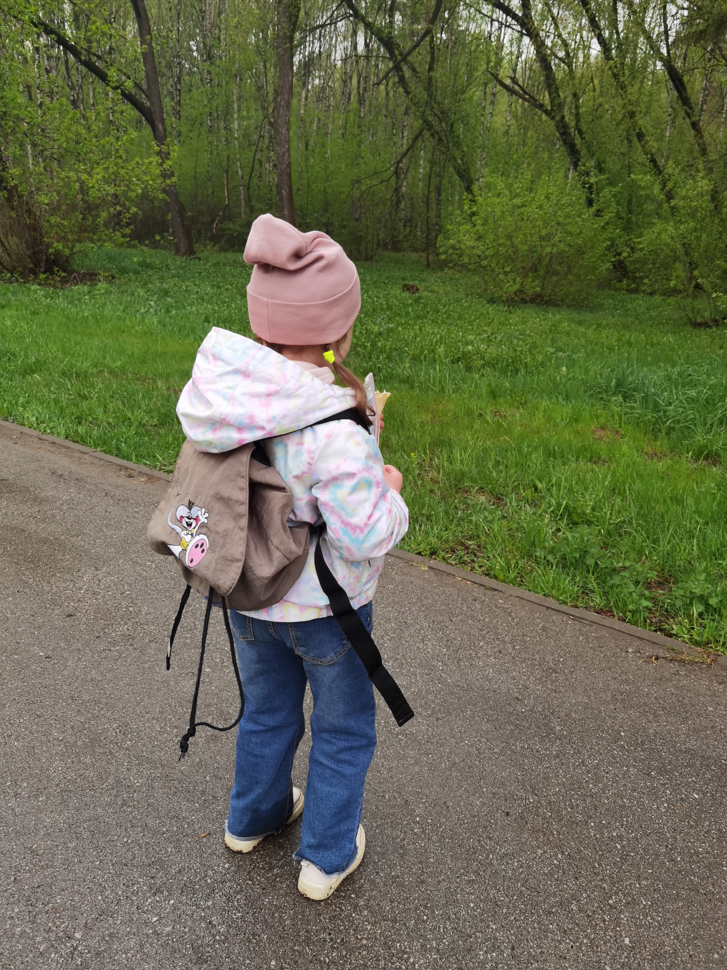A young child, viewed from the rear, stands on a paved surface next to a grassy area bordered by trees, likely a park. The child wears a pink beanie, a light-colored hooded jacket with a patterned design, blue jeans, and white sneakers. A tan backpack with a cartoon character is slung over their shoulders. The child holds something in their hands and looks toward the grassy area. The location is possibly in a park within the city of Perm, Russia, given the presence of trees and green space.
