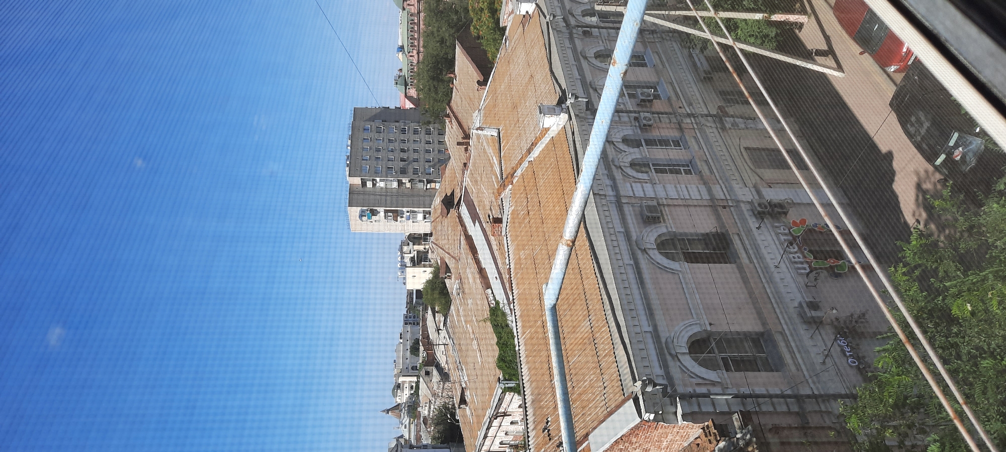 The image captures a view of the city of Astrakhan, Russia, looking towards the northeast. In the foreground, a building with a pinkish-gray facade and arched windows is visible, with a corrugated metal roof that has oxidized to a rusty brown color. A light blue metal pipe runs horizontally across the frame in front of this building. Above and beyond, more buildings with similar roofs and various architectural styles are spread across the cityscape, including a taller, grey apartment building with multiple balconies. Trees and greenery dot the landscape, interspersed between the structures. The sky above is a clear, bright blue with faint white clouds. The scene is observed through a screen, indicated by the distinct horizontal lines overlaying the image. The overall impression is of an urban environment under a clear, sunny sky.