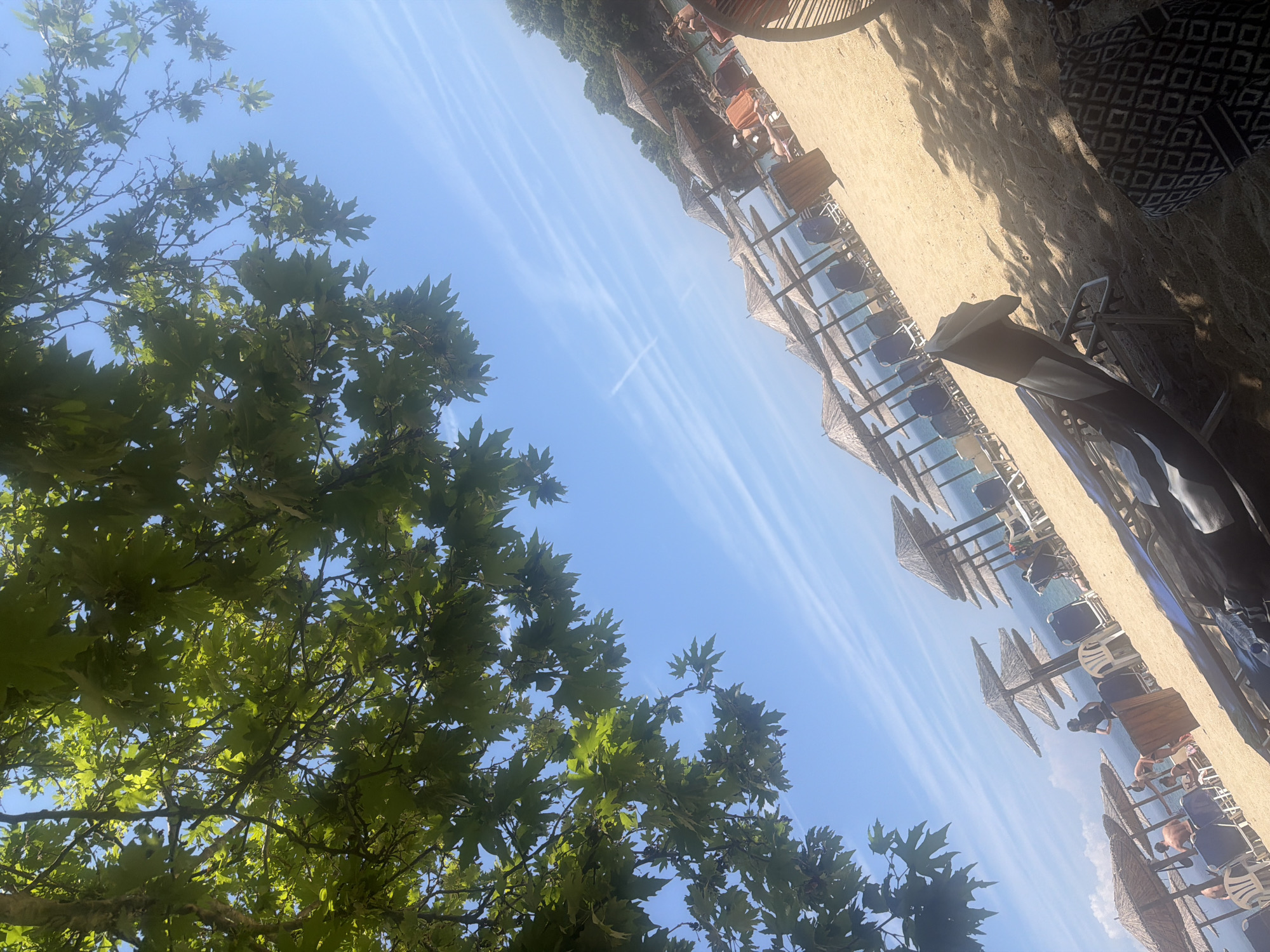 The image depicts a sunlit beach scene in Ολυμπιάδα, Greece, viewed from beneath dense green tree foliage. A bright blue sky dominates the upper right, featuring several faint, linear white contrails. Approximately fifteen to twenty straw-thatched beach umbrellas are regularly spaced along the sandy shore, each accompanied by several dark blue or white lounge chairs. The light-colored sand shows distinct patterns of sun and shadow. The calm, blue Aegean Sea is visible beyond the line of umbrellas. On the right, a densely forested hillside slopes down to the water. In the mid-ground, a person wearing a dark top stands near a cylindrical wooden waste receptacle. Several other individuals are visible sitting on lounge chairs under umbrellas. In the immediate foreground, partially obscured by shadow, a dark lounge chair and a patterned black and white bag or towel are present, along with the arm and shoulder of a person seated in a chair on the far right.