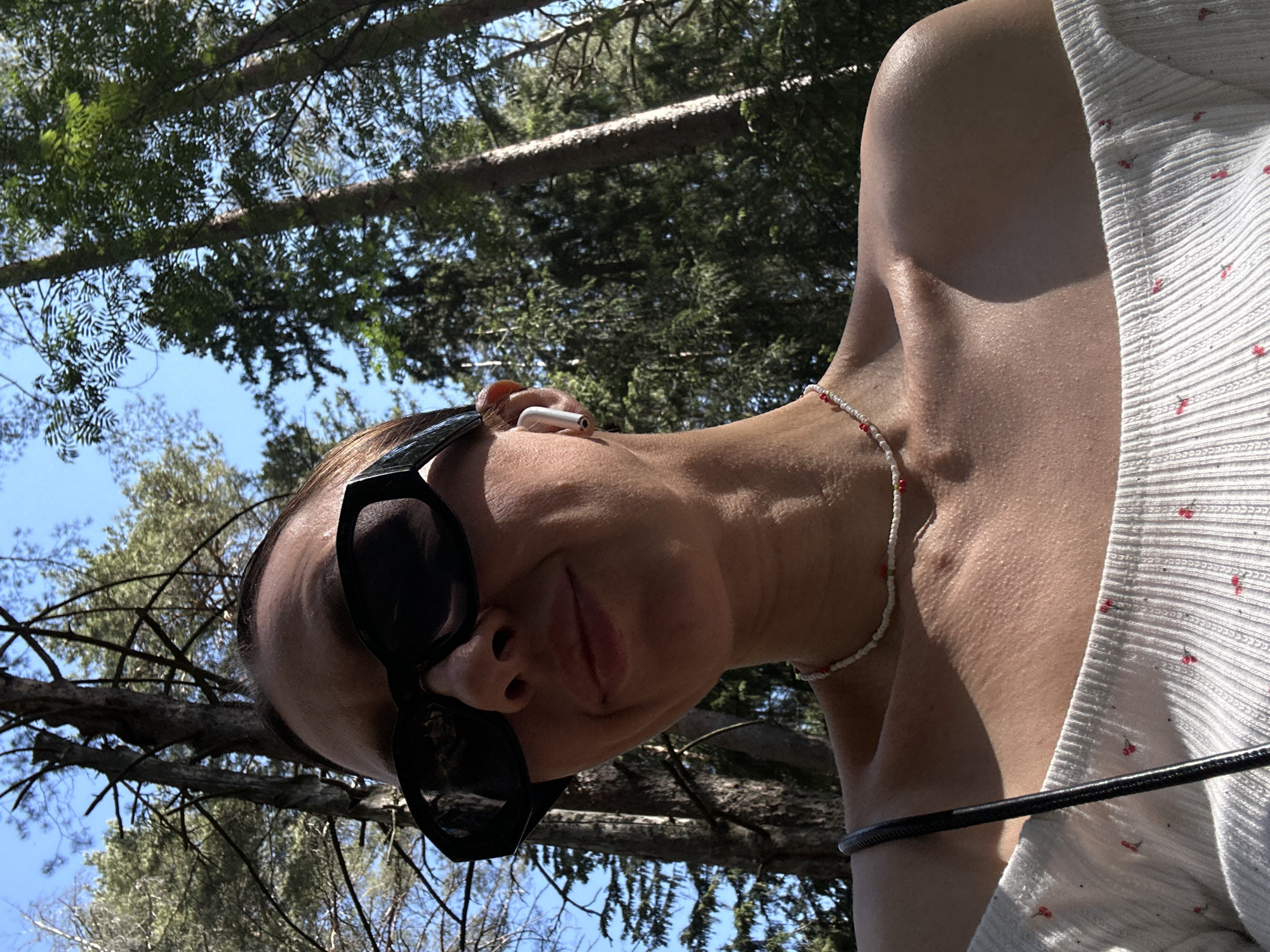 The photograph is taken from a low-angle perspective, looking up at a person with sunglasses and a beaded necklace. The person has an earbud in their ear and is wearing a light-colored shirt with a small pattern. In the background are trees and a blue sky. The specific location in Votkinsk, Russia cannot be determined from the provided image.