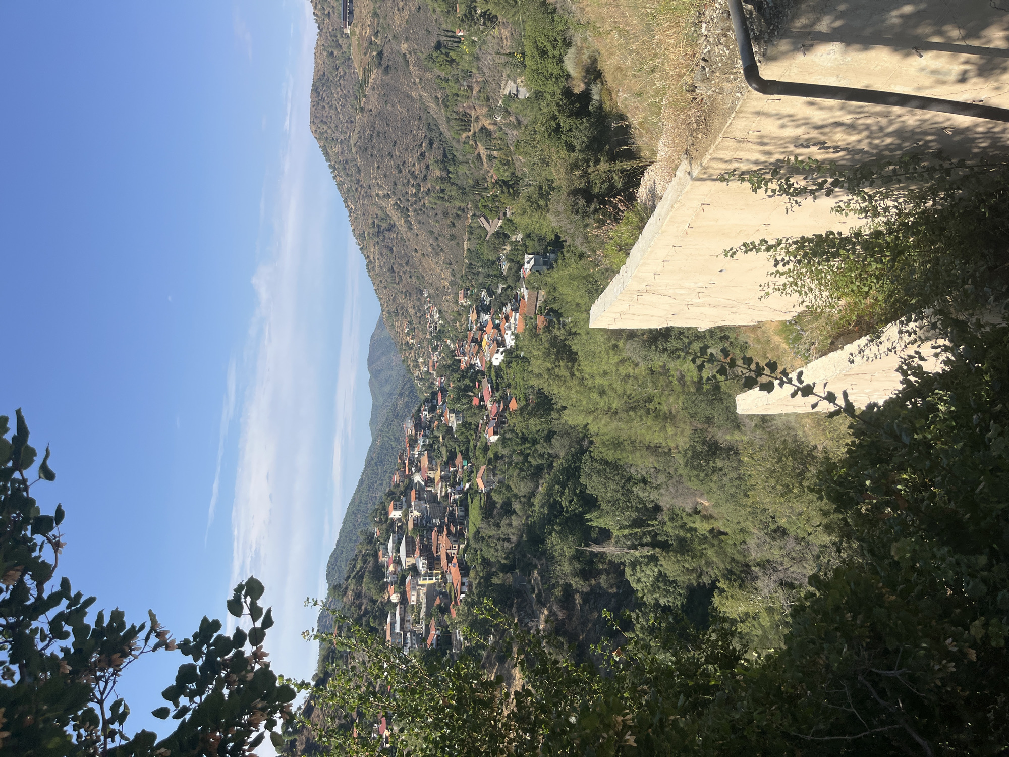 The image shows a village nestled in a valley on a mountainside, identified as Kalopanagiotis, Cyprus. The village features buildings with red-tiled roofs clustered together. The surrounding landscape is hilly and covered with green trees and dry grass, indicative of a Mediterranean environment. In the foreground, a concrete structure with a rough texture and visible rebar is present, casting shadows on the ground. Lush green foliage, including branches and leaves, frames the left side of the scene. The sky is blue with sparse white clouds.