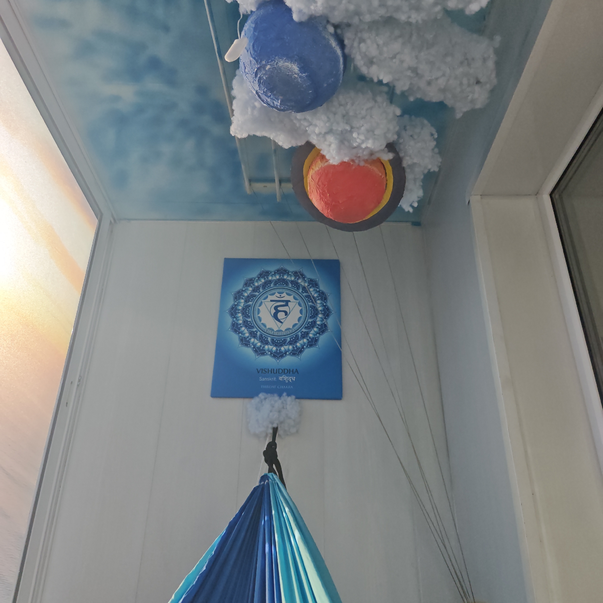 The image shows an indoor corner setting, possibly a balcony, with decorative elements. The ceiling is painted with a sky pattern and features hanging planets surrounded by cotton-like clouds. A blue square card with an intricate circular design labeled "VISHUDDHA" and "THROAT CHAKRA" hangs on the wall. Below the card is a blue hammock suspended by cords. The location is potentially Rostov, Russia, but this is not definitively confirmed by the image.