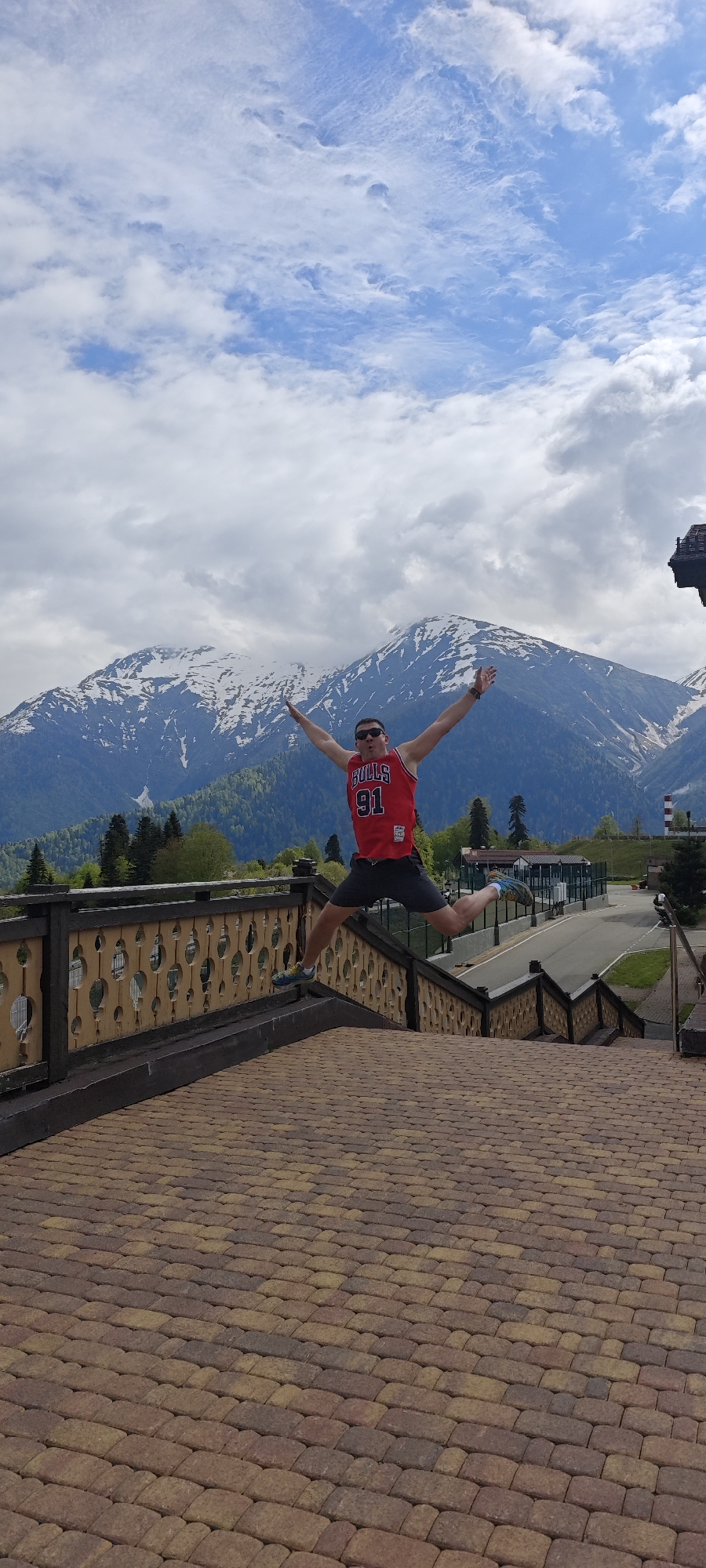 A person in a red basketball jersey is leaping in the air with arms and legs outstretched. The jersey has the word "BULLS" and the number "91" on it. The person is wearing sunglasses and dark shorts. They are positioned above a brick-paved area, with a railing and a descending staircase behind them.
In the background, there are mountains with snow on their peaks. Green trees are visible in the lower part of the mountain range. There is also a road and some buildings in the distance, along with a smokestack. The sky above is cloudy with patches of blue.
The location is identifiable as Rosa Khutor in the city of Unknown, Russia, based on the landscape and architectural elements.