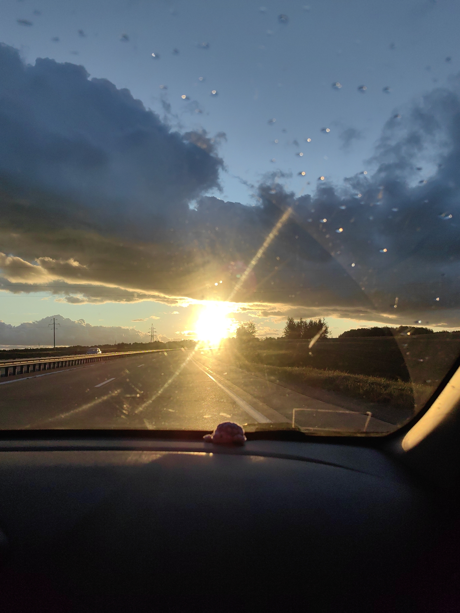 The image is a first-person perspective from inside a vehicle driving on a highway. The sun is setting or rising, casting a warm, golden glow across the scene and creating lens flare effects on the windshield. Dark, dramatic clouds are present in the sky, contrasting with lighter clouds illuminated by the sun. Water droplets are visible on the windshield. The highway stretches forward, with a barrier on the left and fields and trees on the right. Power lines and poles are visible in the distance. The dashboard of the car is in the foreground, with a small, pink decorative item on it. The location is specified as Faniapal'ski sel'ski Savet, Belarus.