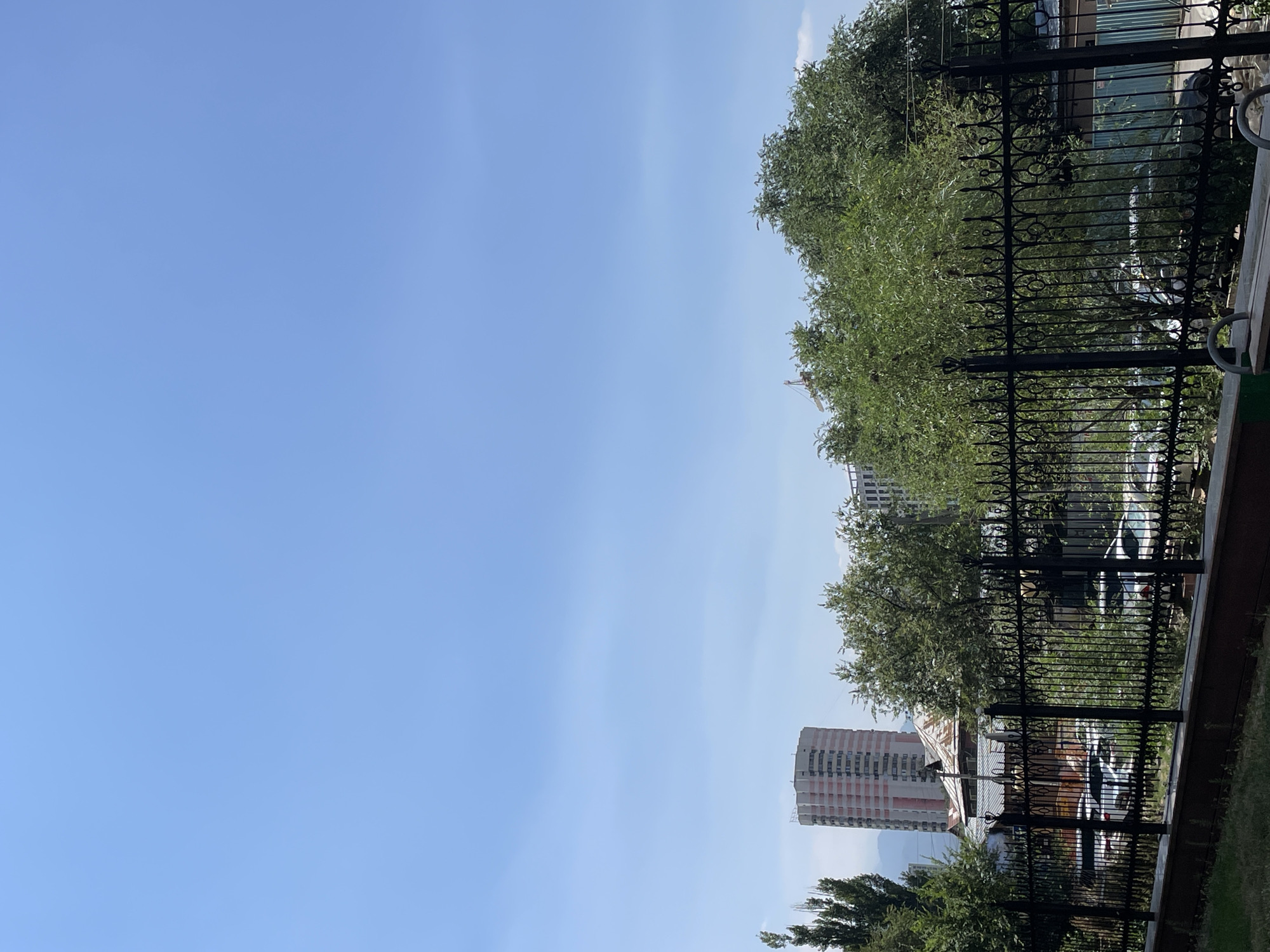 This is a photograph of an urban scene in Almaty, Kazakhstan, taken on a clear, bright day. The foreground is dominated by a black, ornate metal fence. Beyond the fence, several cars are parked amidst lush green trees and bushes. In the mid-ground, a traditional-looking house with a corrugated metal roof is visible. In the background, a tall, modern residential building with a pink and white facade stands out against the clear blue sky. To the far left, a narrower, taller building is also visible. Further in the distance, hazy mountains can be seen. The overall impression is that of a residential area with a mix of older and newer architecture, bordered by greenery.