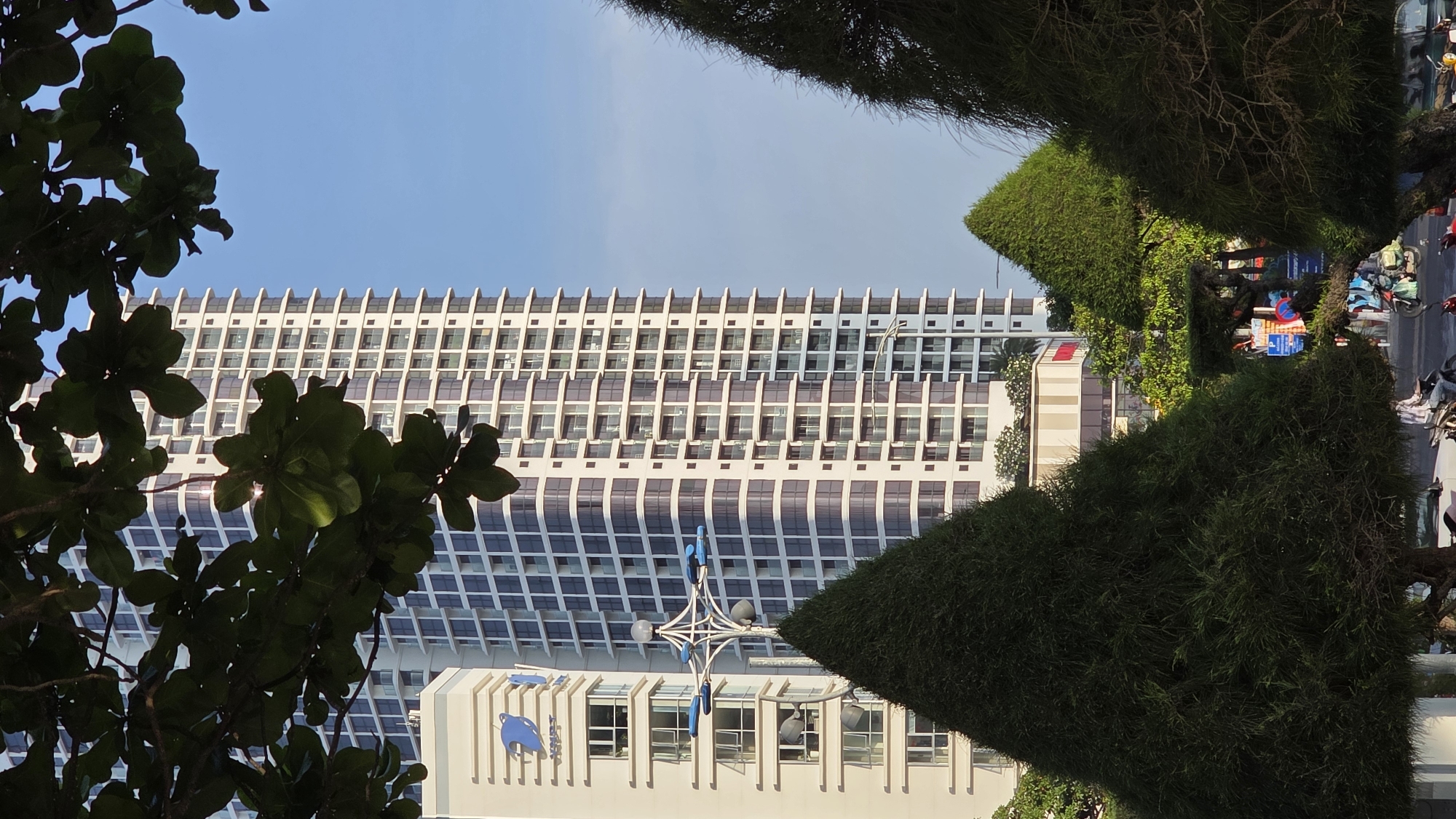 The image displays an urban street scene in Nha Trang, Vietnam, under a bright, clear blue sky. A prominent, modern, and tall building with a distinctive facade dominates the background. Its exterior features a repeating pattern of horizontal and diagonal elements, likely architectural sunshades or decorative panels, with numerous windows reflecting the light.
In the midground, a lower white building is visible, featuring a blue, stylized wave-like logo or emblem on its facade. In front of this building stands a decorative street light structure, painted in white and blue, with multiple arms and light fixtures.
The foreground and right side of the image are framed by dense, green vegetation. Dark leaves from a tree are visible close to the camera on the left, while neatly trimmed, thick bushes or hedges line what appears to be a park or street median, partially obscuring the view of the street. Beyond these bushes, a bustling street scene unfolds, with several motorbikes parked or in motion, and indistinct figures of people, suggesting everyday urban activity. A distant building with a red sign is partially visible on the far right.
The lighting indicates daytime, likely morning or late afternoon, with bright, direct sunlight illuminating the scene. The weather is sunny with no clouds. The architectural style, the specific street furniture, and the presence of numerous motorbikes are typical characteristics of urban environments in Vietnam. The blue logo on the building serves as a unique location cue. No clearly legible text is visible beyond the blue logo.