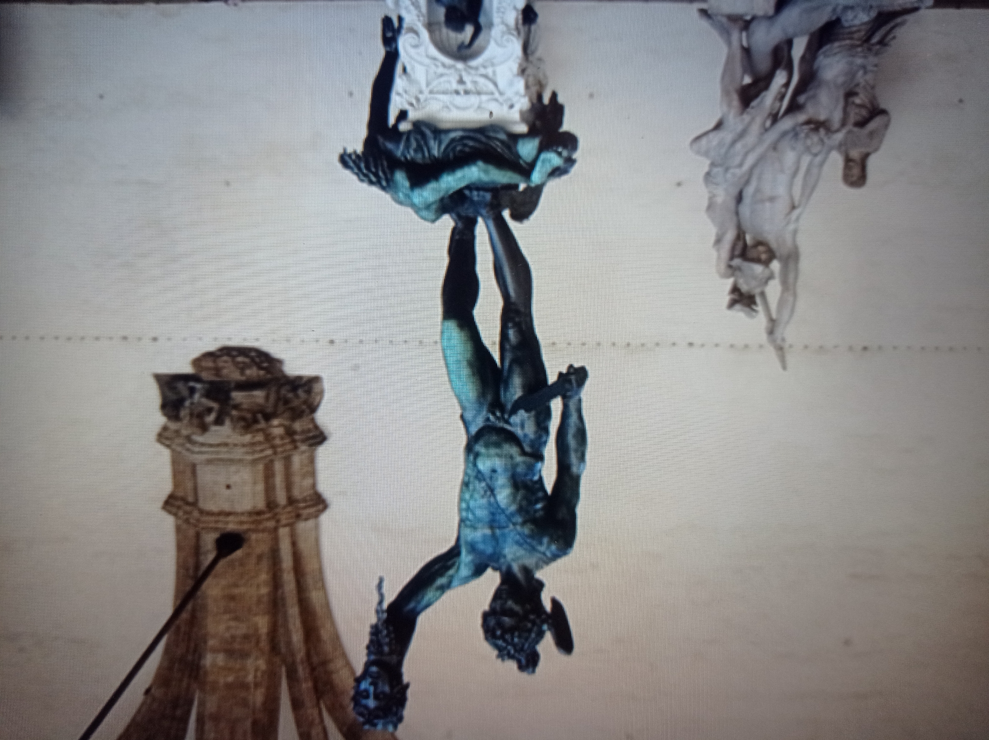 The image shows a bronze sculpture of Perseus holding the severed head of Medusa. He stands triumphantly on the body of Medusa, with a sword in one hand and Medusa's head in the other. To the left of the main sculpture, a lighter-colored marble sculpture depicts another mythological scene with several figures. In the background, a portion of a decorative architectural element, possibly a column or archway, is visible. The sculptures are placed against a plain, textured wall. The scene is a public art display. The location is not in Mukachevo, Ukraine.