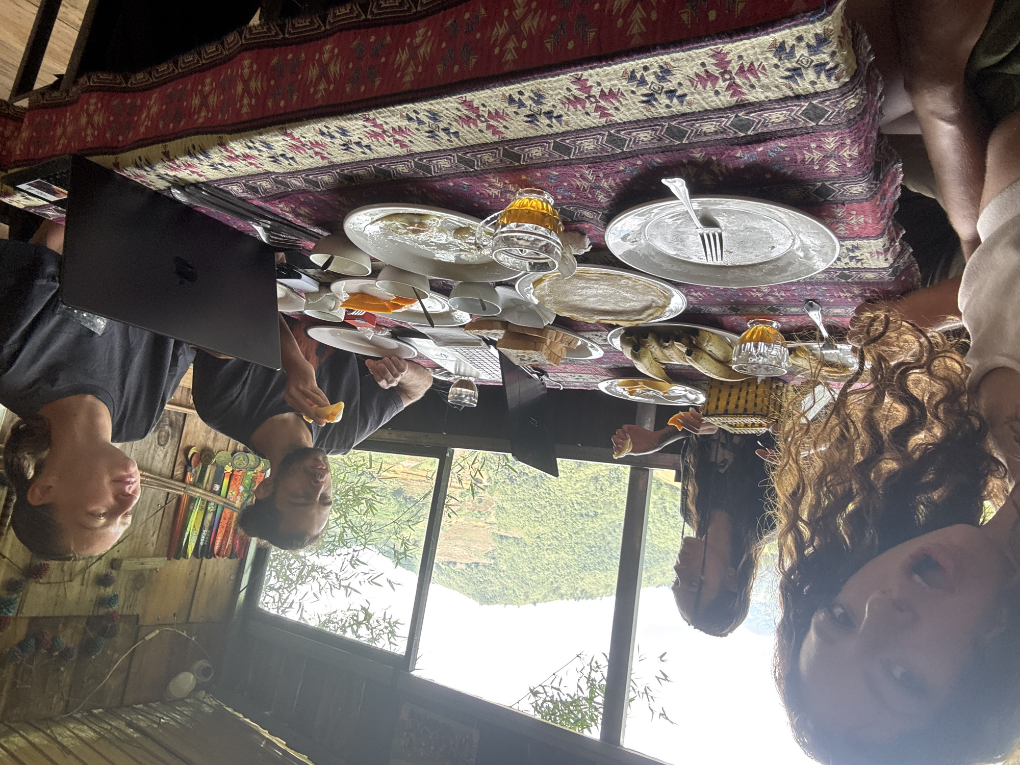 A medium shot, from an upside-down perspective, reveals four individuals seated around a table laden with dishes. The individuals appear to be engaged in a meal. A laptop is placed on the table, suggesting a blend of work and leisure. The location is in Ngã Ba Kim, Vietnam, as suggested by the mountain range and landscape visible through the windows. The room appears to be a rustic setting with wooden walls. The table is covered with a patterned cloth, and plates, cups, and remnants of food are scattered across its surface. Each person is holding a piece of food.