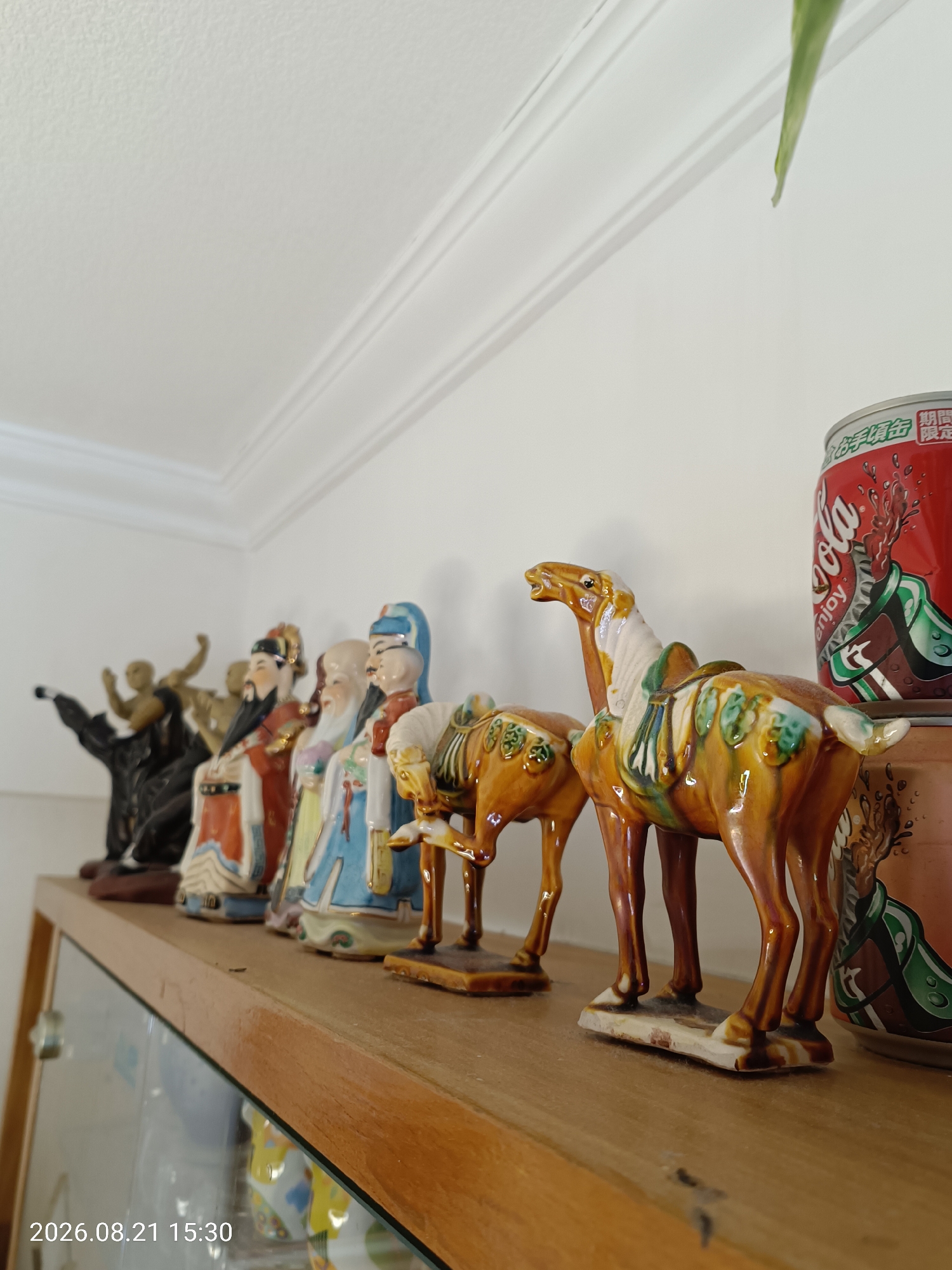 A close-up, low-angle shot captures a collection of ceramic figurines and a soda can displayed on a wooden shelf. The figurines, positioned from left to right, depict several human figures dressed in traditional attire, followed by two horse figurines with saddles. The colors of the figurines are vibrant, with prominent use of brown, yellow, blue, and red. Behind the figurines, a red Coca-Cola can with Asian characters is visible, partially obscured by a larger cylindrical object with green detailing. The background consists of a plain white wall with decorative molding at the top. The lighting is soft, casting gentle shadows. The timestamp "2026.08.21 15:30" is superimposed on the bottom left of the image. The scene appears to be indoors, likely in a residential setting.