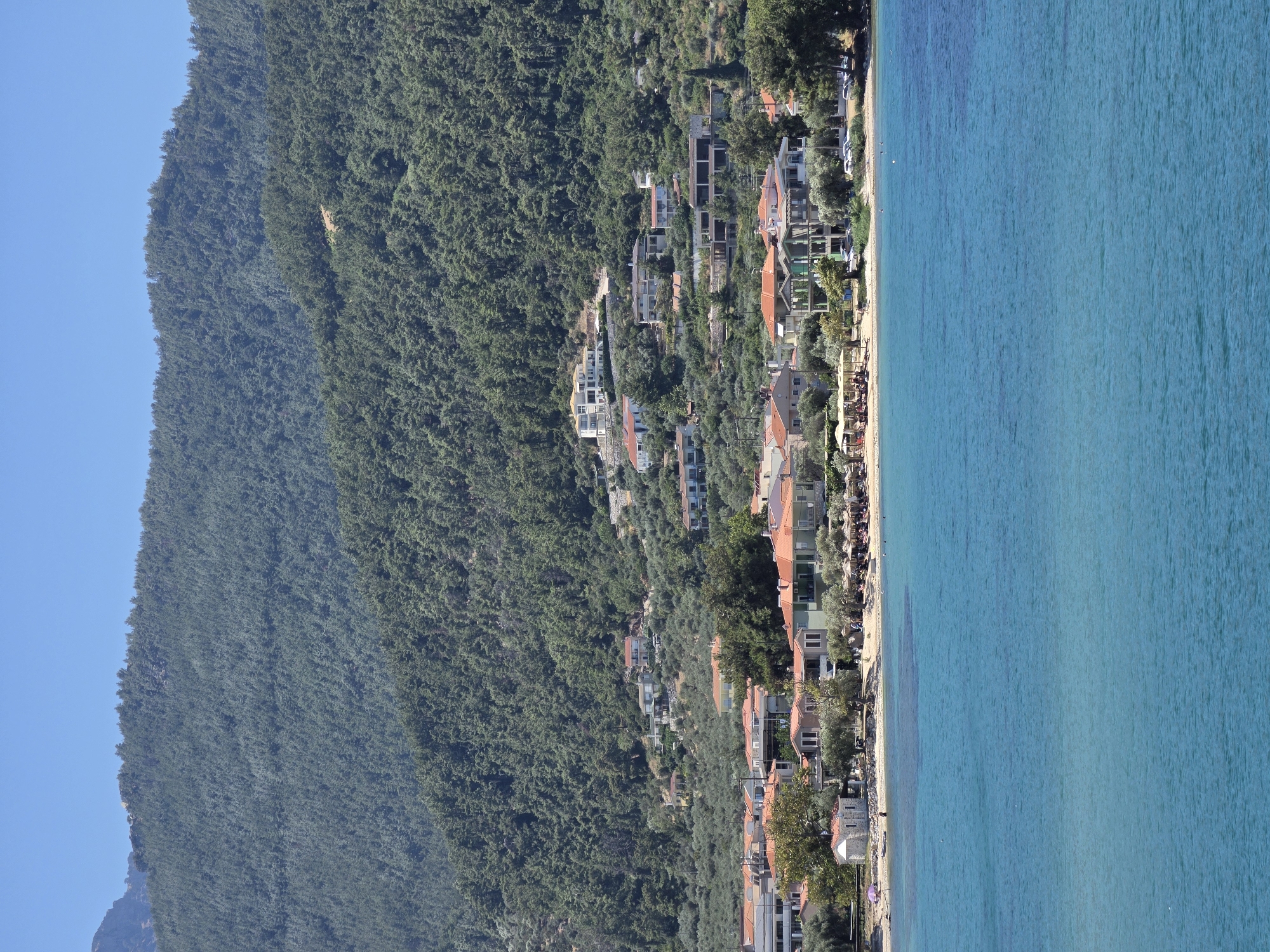 The image displays a coastal town nestled at the base of a densely forested mountain. The foreground is dominated by the tranquil, clear turquoise waters of the sea. The middle ground features a sandy beach lined with buildings, some with orange tile roofs, and numerous umbrellas providing shade. A few individuals are visible on the beach, some seated under the umbrellas, suggesting a relaxed atmosphere. Beyond the beach, the mountain rises steeply, covered in a thick carpet of green trees, with a few scattered houses visible on its slopes, indicating development on the hillside. The sky above is a clear, bright blue, suggesting a sunny day. The scene evokes a sense of a peaceful seaside village surrounded by natural beauty.