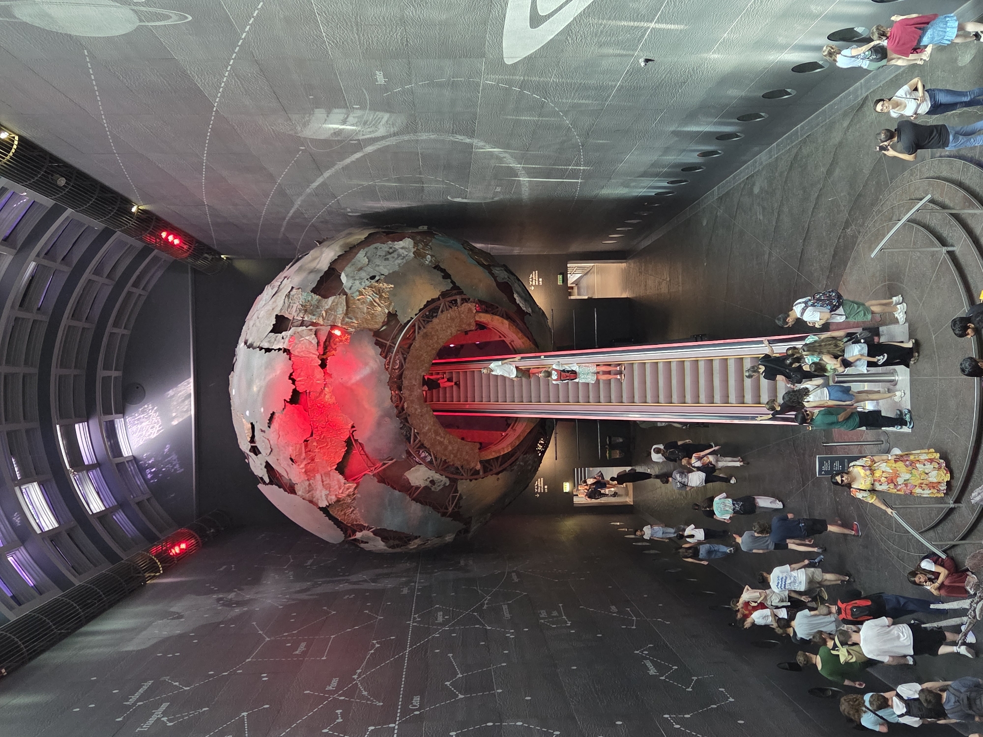 The image shows an interior view of a large, modern space, likely a museum or exhibit hall. The location is the Natural History Museum in the city of London, United Kingdom.
The dominant feature is a large, spherical sculpture that appears to be a representation of the Earth, with exposed sections that reveal its interior structure, illuminated in red. An escalator runs through the center of the sphere.
Numerous people are visible, both on the escalator and walking around the base of the sphere. The floor has a dark surface that is decorated with outlines of constellations and astronomical diagrams.
The ceiling is high and dark, with a pattern of lights and decorative elements. The space appears to be well-lit, though the lighting is primarily focused on the sphere and the people moving around it.