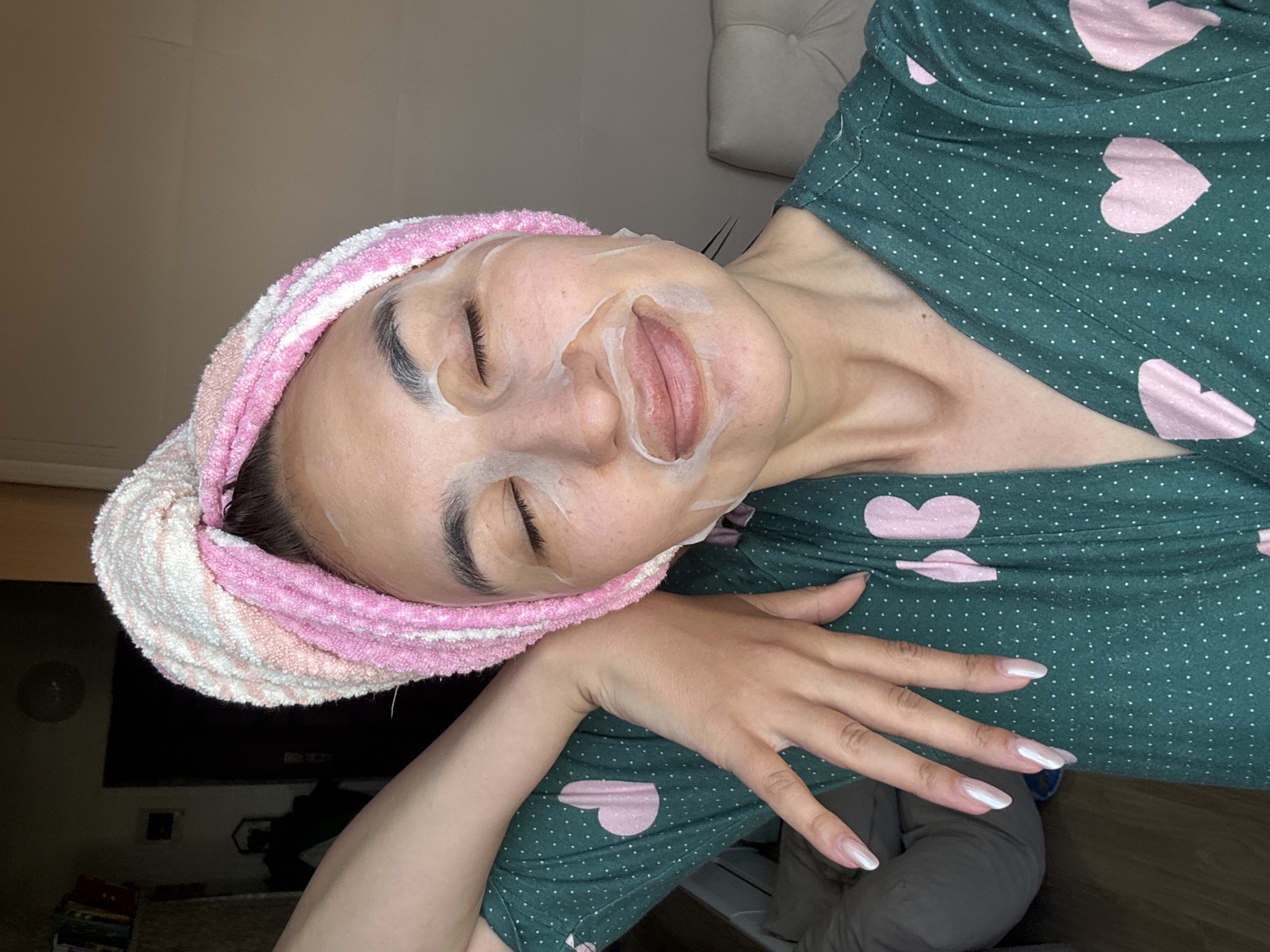 A woman is lying down, wearing a pink and white striped towel wrapped around her head and a green shirt with pink heart patterns. She has a facial mask on her face and her eyes are closed. Her left hand is extended with long, manicured nails. The background shows a neutral-colored wall and some indistinct objects, including a glimpse of a fan, and an area on the floor with some stacked items, indicating an indoor environment. There is nothing in the image to indicate the location is in Sochi, Russia.