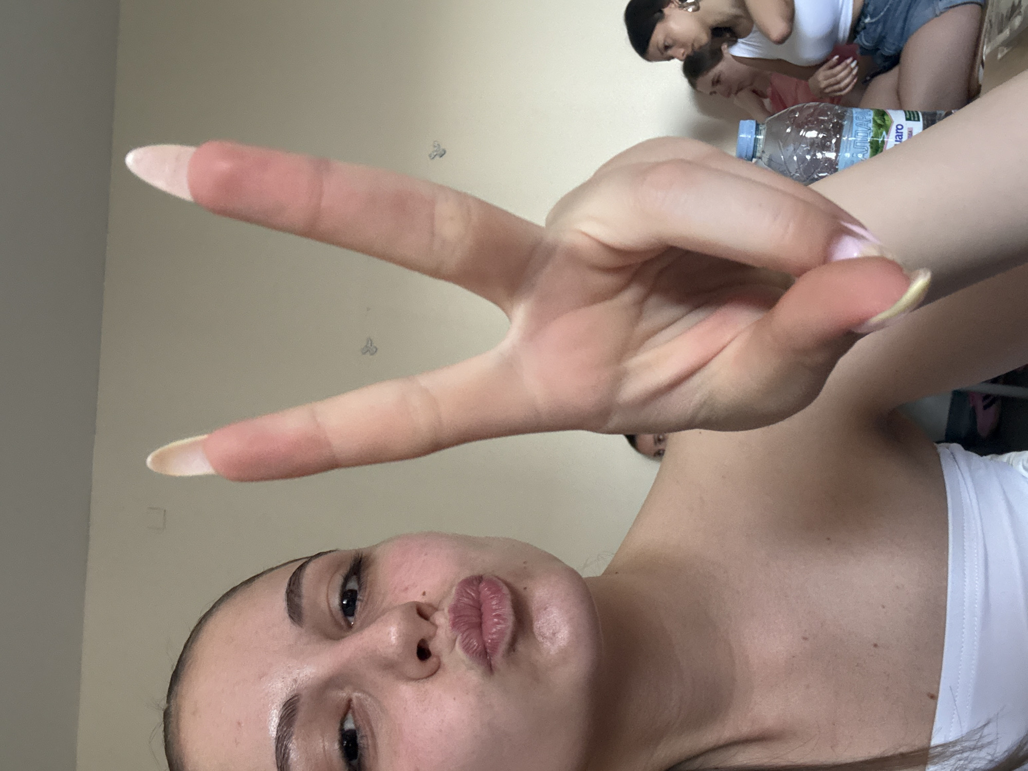 A close-up selfie shows a light-skinned woman with dark hair and eyes puckering her lips and making a "peace" sign with her hand. Her fingernails are long and painted a light color. In the background, there are three other people, a plastic water bottle, and a light-colored wall with what appear to be decorative stickers. It's not possible to determine the exact location in Burgas, Bulgaria, from this image alone.
