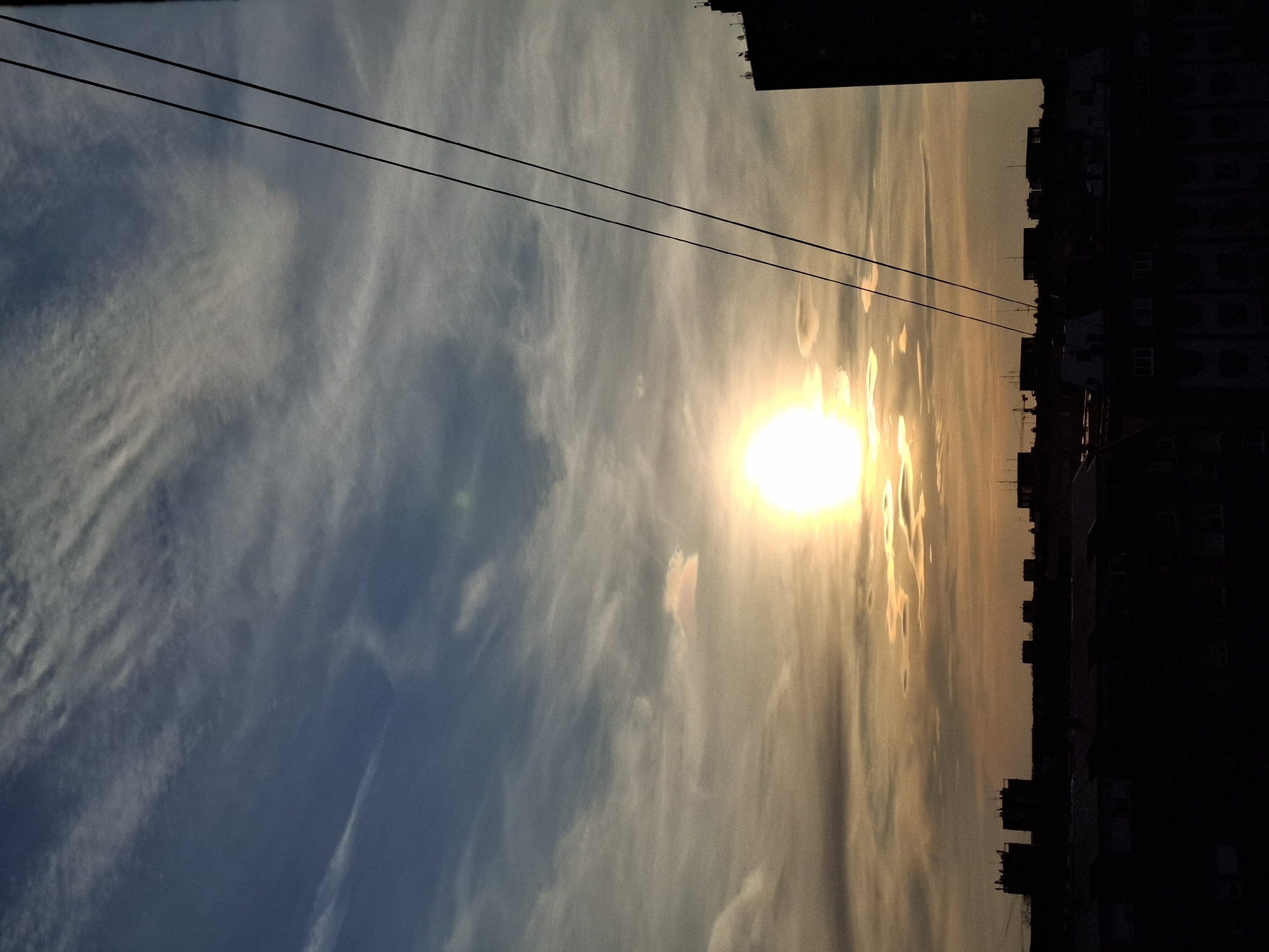 A photograph taken in Novi Sad, Serbia, shows a low-angle perspective of the sky at sunset. The sun, a bright, circular shape, is positioned towards the center-right of the frame. The sky transitions from a lighter, golden hue around the sun to a deeper blue color on the left side of the image. Cirrus clouds are visible throughout the sky. Two parallel power lines stretch across the top portion of the image from left to right. The silhouettes of buildings are visible on the right side, forming a dark, irregular line against the bright sky.