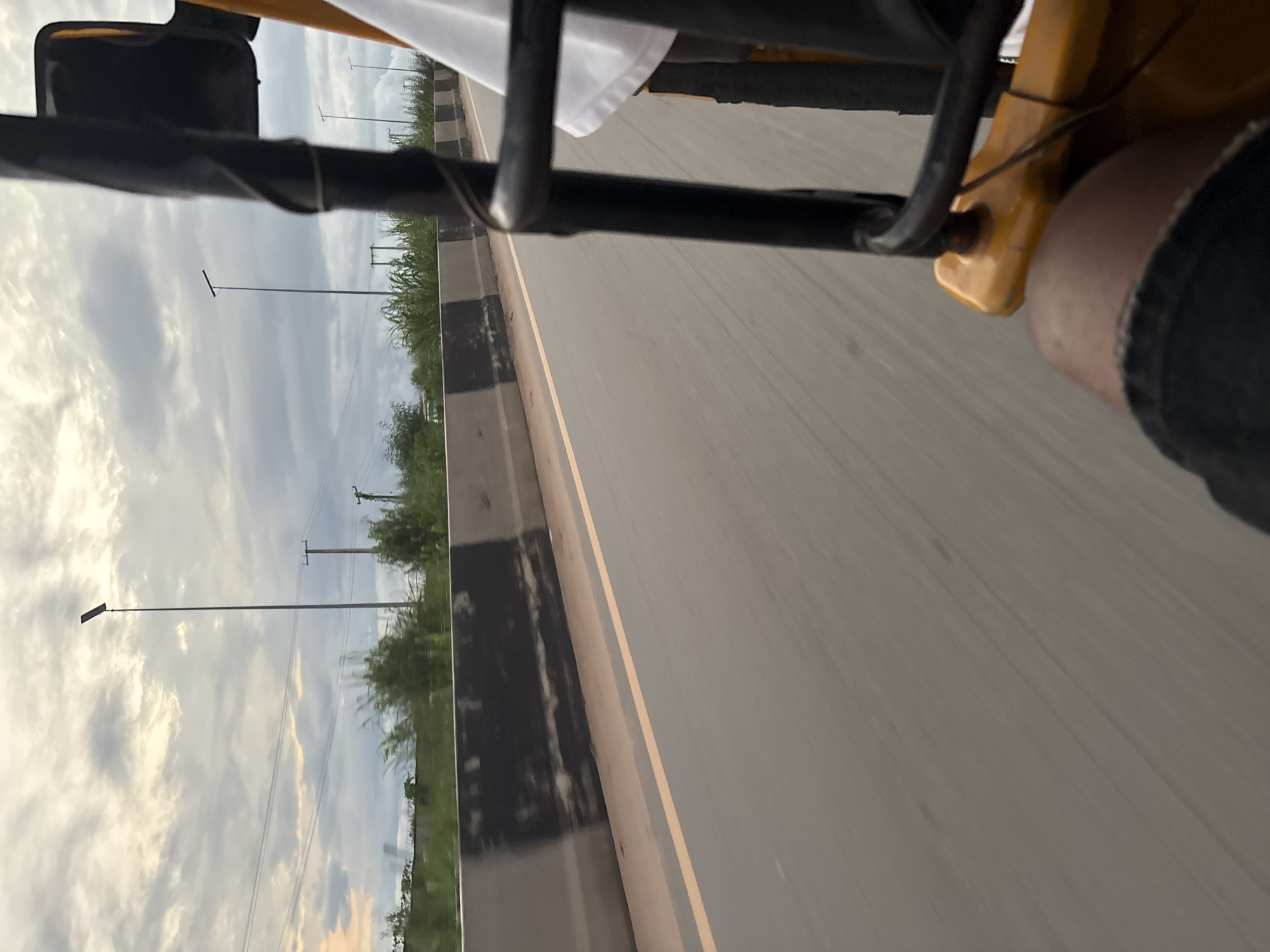 A first-person perspective shot from the back of a yellow tricycle in Onitsha, Nigeria, shows a road with a concrete barrier separating it from vegetation and streetlights in the distance. The sky is cloudy and overcast. The tricycle has a black metal frame and a yellow seat. The driver's leg, clad in dark denim shorts, is visible in the foreground. The tricycle appears to be in motion, indicated by the motion blur on the road.