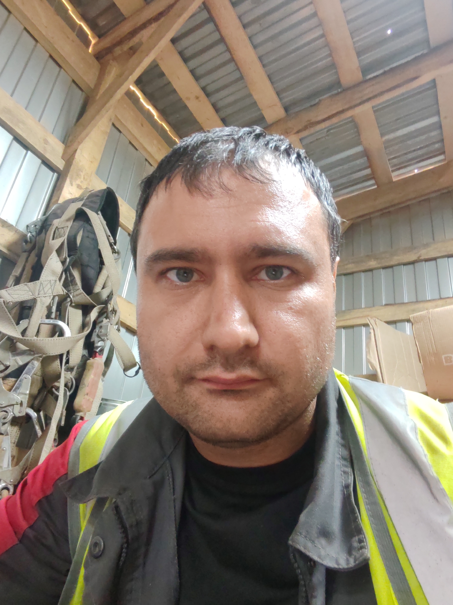 A man is looking at the camera in what appears to be a storage area or workshop. He is wearing a black t-shirt, a grey jacket with red sleeves, and a yellow high-visibility vest over the jacket. To his left, a piece of safety equipment, possibly a harness, is hanging on a wall. The background shows wooden beams, corrugated metal walls, and some cardboard boxes. The lighting is somewhat dim, with reflections visible on the man's forehead, suggesting a warm or humid environment. The scene is likely indoors. There is no specific information provided about the location being in Murashi, Russia.
