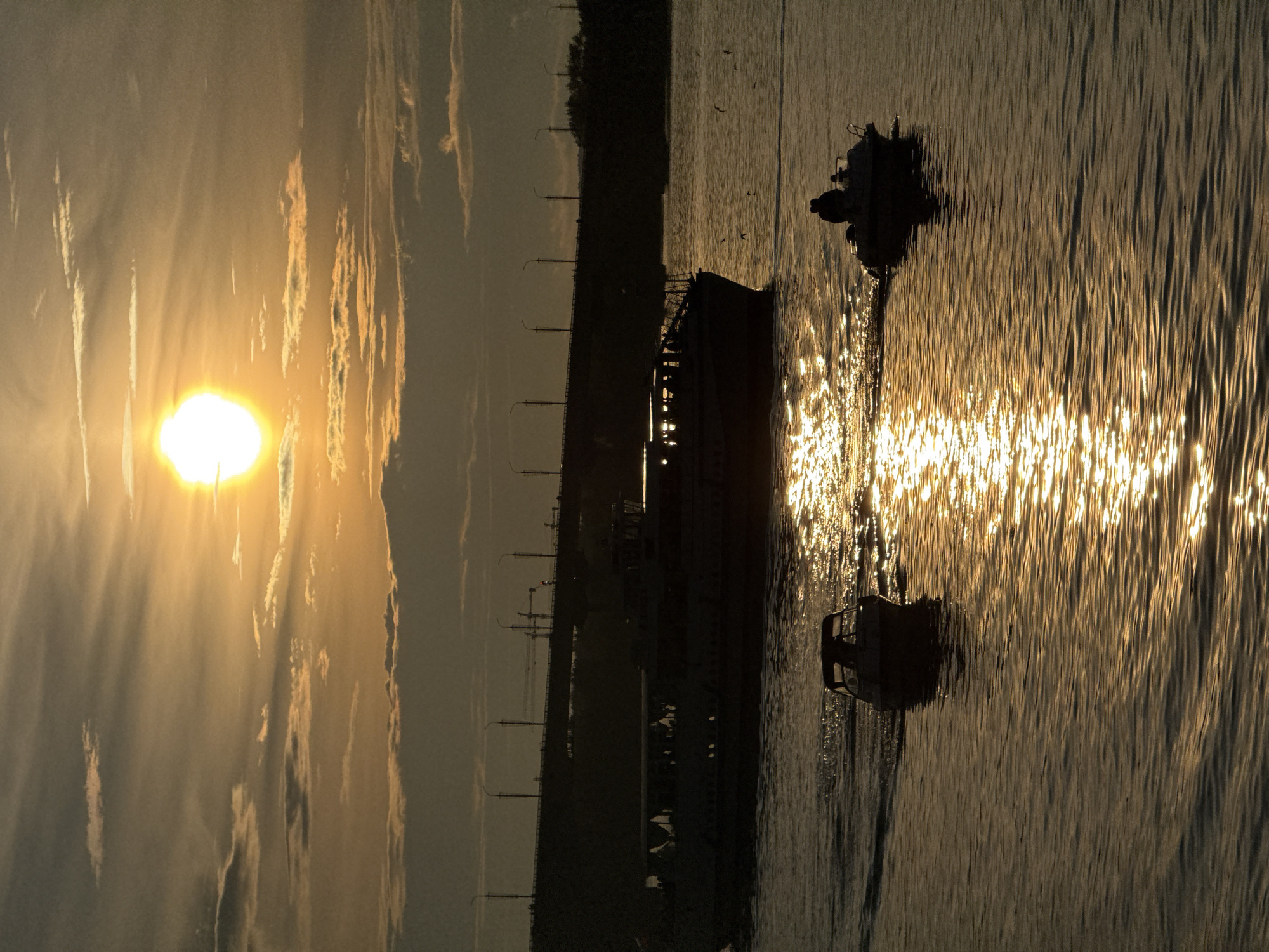The photograph captures a serene sunset over the Kama River in Perm, Russia. The sun, a bright orb, hangs low in the sky, casting a warm, golden glow that reflects off the rippling water. In the foreground, two small boats navigate the river, their silhouettes stark against the shimmering surface. One boat, closer to the viewer, carries a single figure. The other, further out, is occupied by two people. Dominating the mid-ground is a larger passenger boat, also silhouetted, with passengers visible on its deck. In the distance, a bridge spans the river, its lights faintly visible, and a line of streetlights stretches along the embankment. The sky is a canvas of soft clouds, tinged with the hues of the setting sun. The overall atmosphere is one of tranquility and the peaceful close of a day.