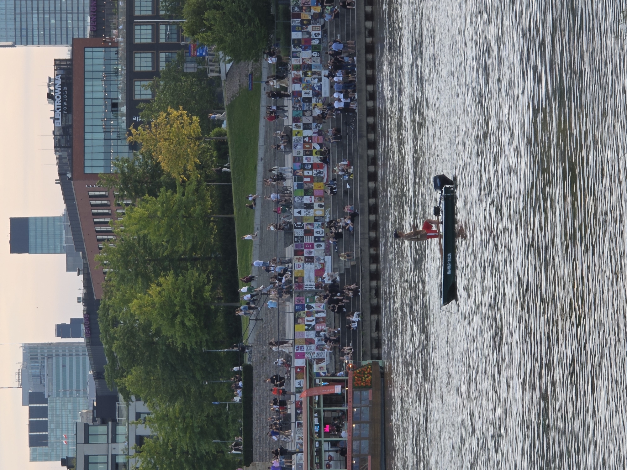 The image depicts a scene along the Vistula River in Warsaw, Poland, with a focus on the Elektrownia Powiśle development. In the foreground, a dark green motorboat with a person standing in it, wearing red shorts and a light-colored shirt, is on the rippling water. The boat has an outboard motor and a registration number, "MA-WAR-11852A," visible on its side.
Behind the river, a wide, tiered embankment is populated with numerous people sitting, standing, and walking. The lower tiers of the embankment are adorned with a vibrant display of posters and artwork. Above the embankment, lush green trees form a canopy, partially obscuring modern buildings and skyscrapers in the background. The most prominent building in the mid-ground is a red brick structure with large glass windows, identified by the illuminated sign "ELEKTROWNIA POWIŚLE". Further in the background, contemporary high-rise buildings, including one with a Polish flag, characterize the Warsaw skyline. A portion of a docked passenger boat with seating and an awning is visible on the left side of the image, near the embankment. The sky is a pale, overcast hue, suggesting it might be dawn or dusk.