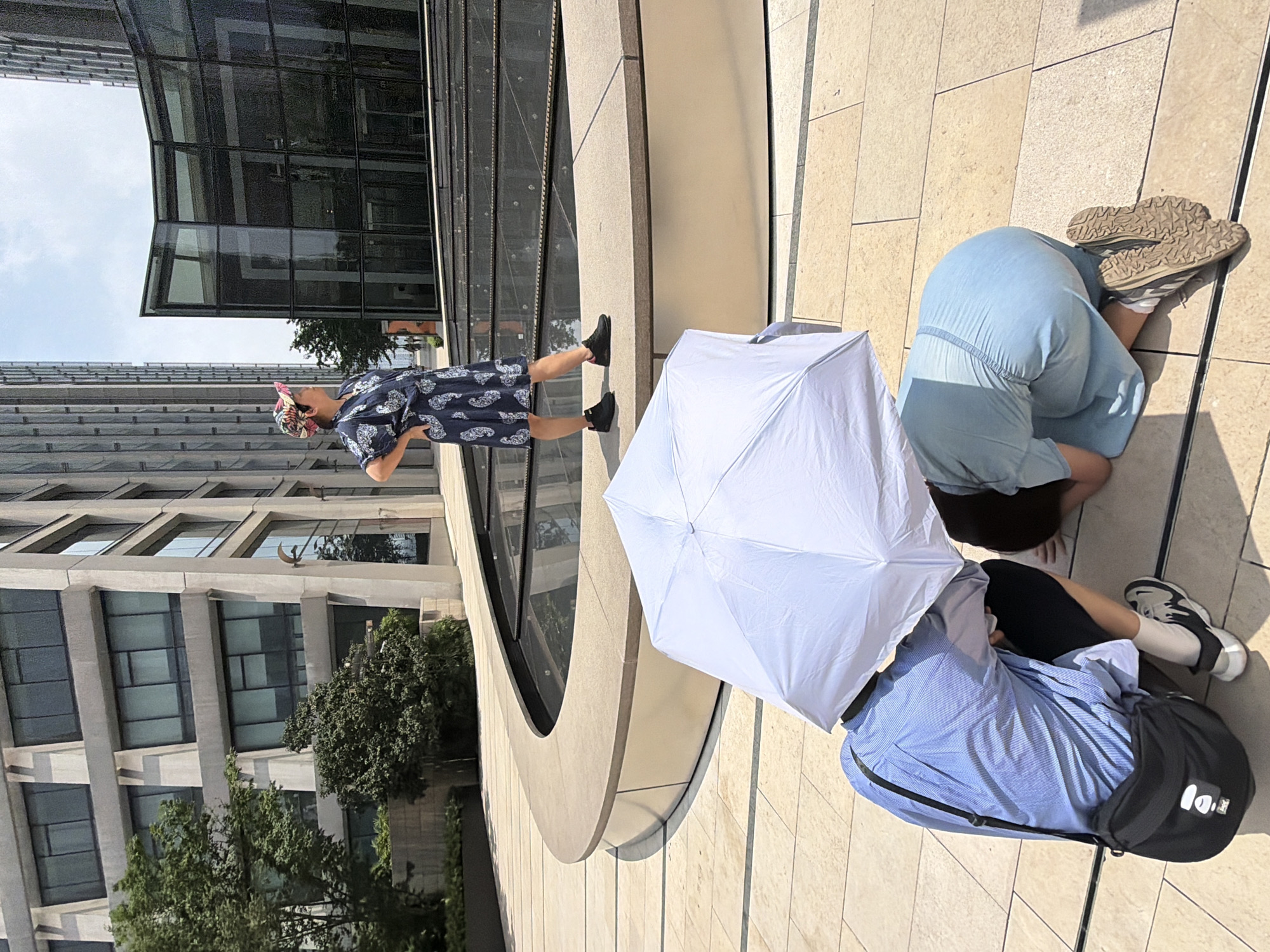 In Yuzhong District, China, a woman stands on a raised concrete platform in front of a modern glass building and a water feature. She wears a patterned dress and a colorful cap, with her hands on her hips. In the foreground, two other individuals are crouching beneath a large white umbrella. One is wearing a blue striped shirt and a black bag, and the other is wearing a light blue dress. The scene appears to be outdoors on a sunny day, with tiled ground and architectural elements of a city environment.