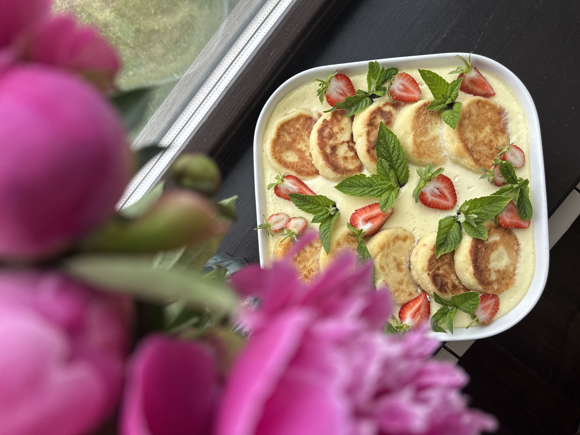 In the city of Chaikovsky, Russia, a close-up shot captures a white rectangular dish filled with syrniki, a type of fried quark pancake, swimming in a creamy yellow sauce. The syrniki are golden brown and round, arranged in a pattern on top of the sauce. Halved strawberries and fresh mint leaves are artfully scattered around the syrniki, garnishing the dish. In the foreground, out of focus, are large pink peony petals, adding a soft, floral element to the scene. The dish rests on a dark, textured surface, possibly a wooden table. In the background, a windowsill with a white frame and a hint of green foliage outside can be seen. The lighting suggests it is daytime.