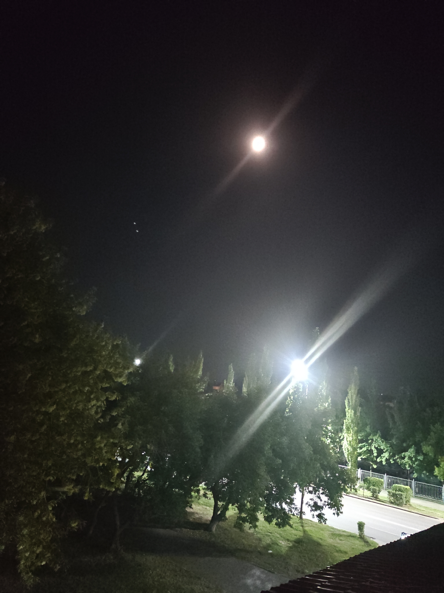 An outdoor night scene at eye-level, likely taken from a rooftop or elevated vantage point. The image is dominated by darkness with bright artificial lights creating streaks and a halo effect. A prominent, bright light source, likely a streetlamp, is positioned in the mid-right of the frame, casting a strong glare and illuminating the surrounding trees and vegetation. In the upper part of the sky, a small, bright, circular object is visible, possibly the moon. Several smaller, pinpoint lights are scattered in the sky to the left of the main light source. In the foreground, the dark roof tiles of a building are visible. Beyond the light source, a road is visible with a white metal fence and manicured bushes lining one side. Trees of various sizes and densities fill the mid-ground, some with illuminated foliage. The overall impression is that of a suburban or park-like area within a city at night, with artificial illumination significantly impacting the visibility of natural elements. The location is specified as Petropavl, Kazakhstan.