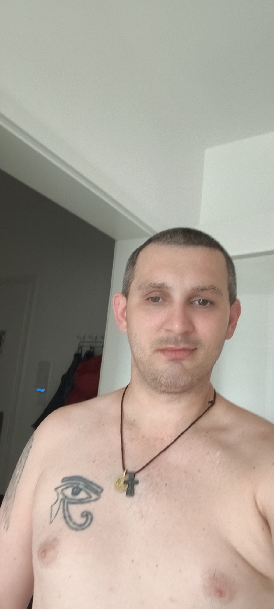 A man with short brown hair and a light complexion is pictured from the chest up, facing the camera directly. He has stubble on his face and is not wearing a shirt, revealing a tattoo of an Egyptian eye on his left chest. He is also wearing a dark cord necklace with a cross pendant and a small circular charm. The background is a plain, light-colored wall with a doorway visible to the left, which leads to a darker interior space where clothing items are hanging on a rack. The lighting appears to be natural, coming from the right. The image was taken in Wolgast, Germany.