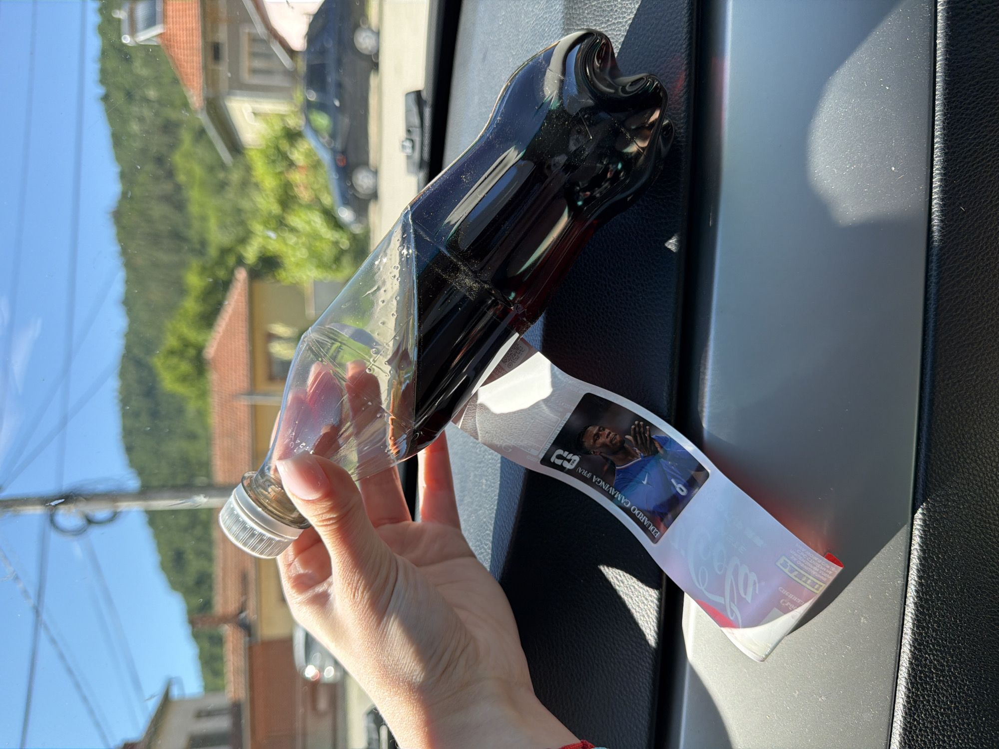 A hand holds a plastic bottle of dark soda, tilted to the right. A sticker with the image of a soccer player named Eduardo Camavinga is attached to the bottle. The bottle rests on a dark surface, likely the dashboard of a vehicle, in front of a blurred background of houses and trees under a bright, sunny sky. The scene appears to be in Beli Osam, Bulgaria, indicated by the context of the image.
