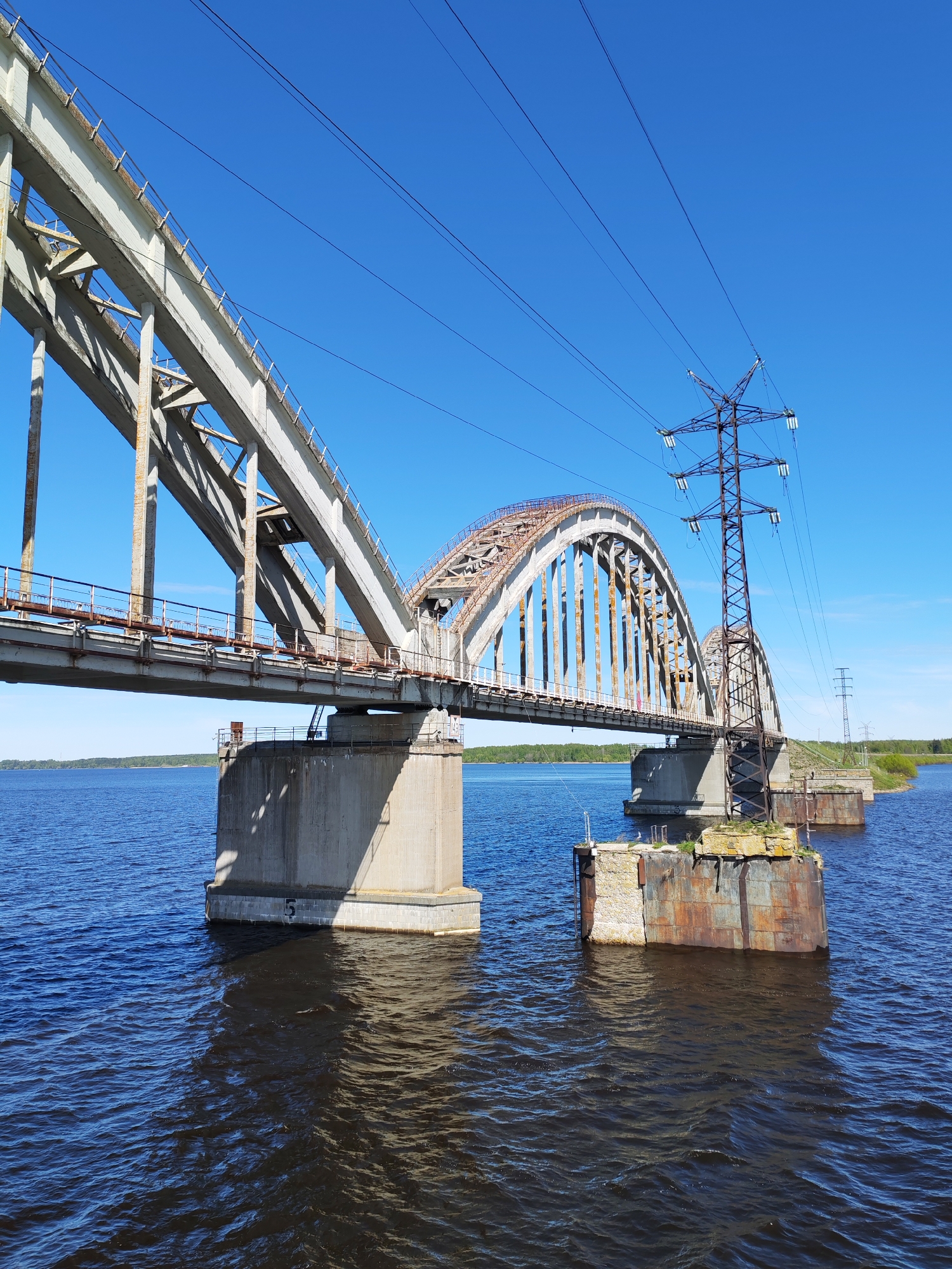 The image captures a bridge spanning a body of water under a clear blue sky. The bridge is primarily constructed of concrete and steel, with a combination of straight sections and an arched portion. A utility pole with power lines is attached to the bridge structure. The water is dark blue and slightly choppy. In the background, a distant treeline is visible along the horizon. The number "5" is painted on one of the bridge's concrete supports. The bridge is located in Gladyshevo, Russia.