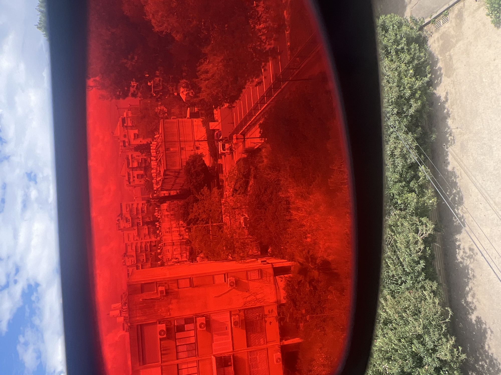 The image shows a view through a red-tinted lens, likely a telescope or binoculars, of a city scene. In the left portion of the frame, blue sky with white clouds is visible outside the lens. Through the red lens, a residential area with multiple buildings of varying heights is seen. Many buildings have air conditioning units. A bridge or walkway is visible amidst trees and other foliage. On the right edge of the frame, outside of the lens, is a concrete area with green bushes and power lines. The location is identified as Kiryat Bialik, Israel.