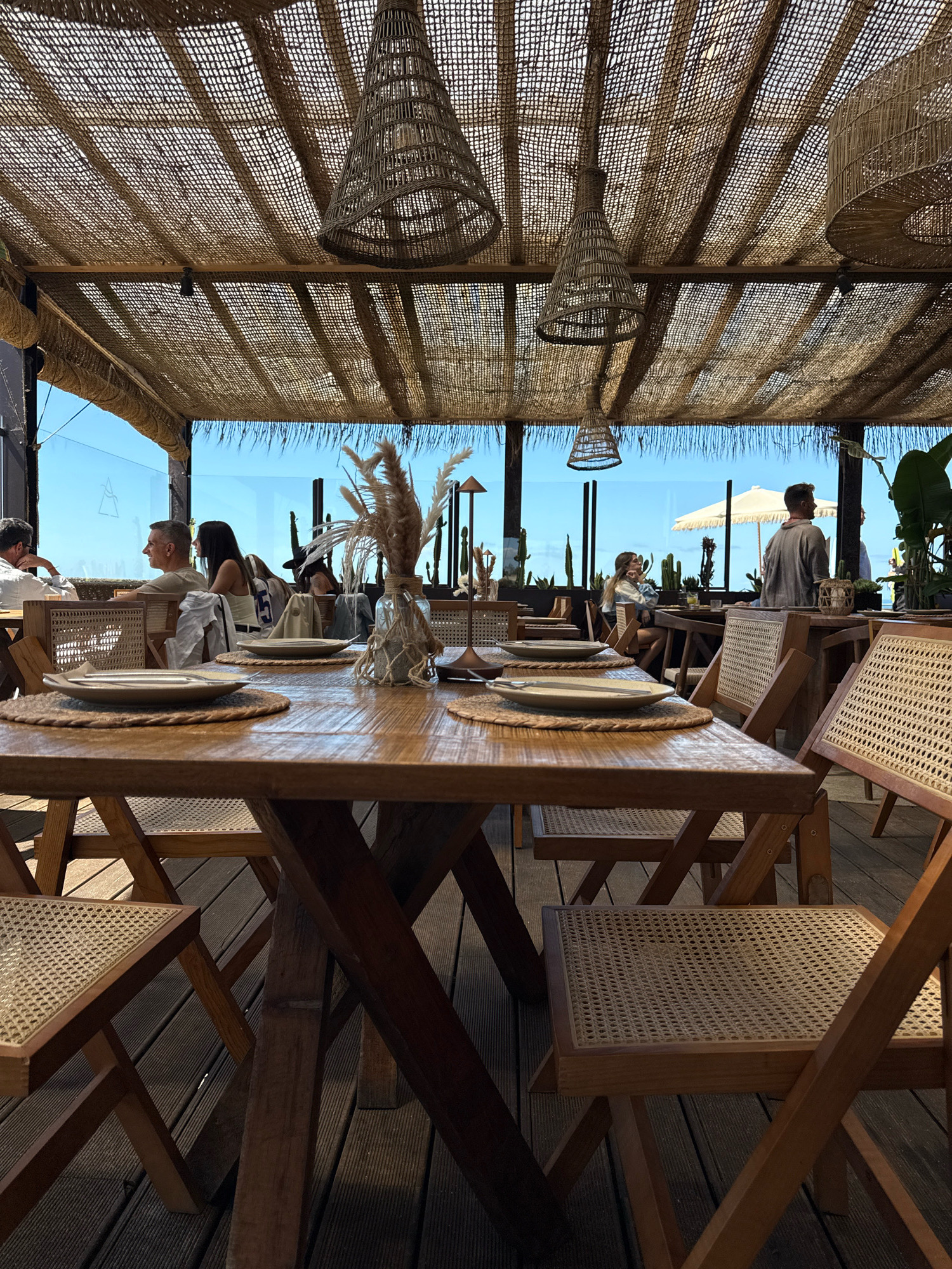The image shows an outdoor dining area with several wooden tables and chairs. The tables are set with plates and placemats. A woven canopy covers the area, with hanging woven lampshades. In the background, people are seated at tables, and there is a view of the sky and some greenery. This scene is likely from a restaurant in Vila Nova de Gaia, Portugal, given the style of the furniture and the open-air setup, common in coastal establishments in the region.