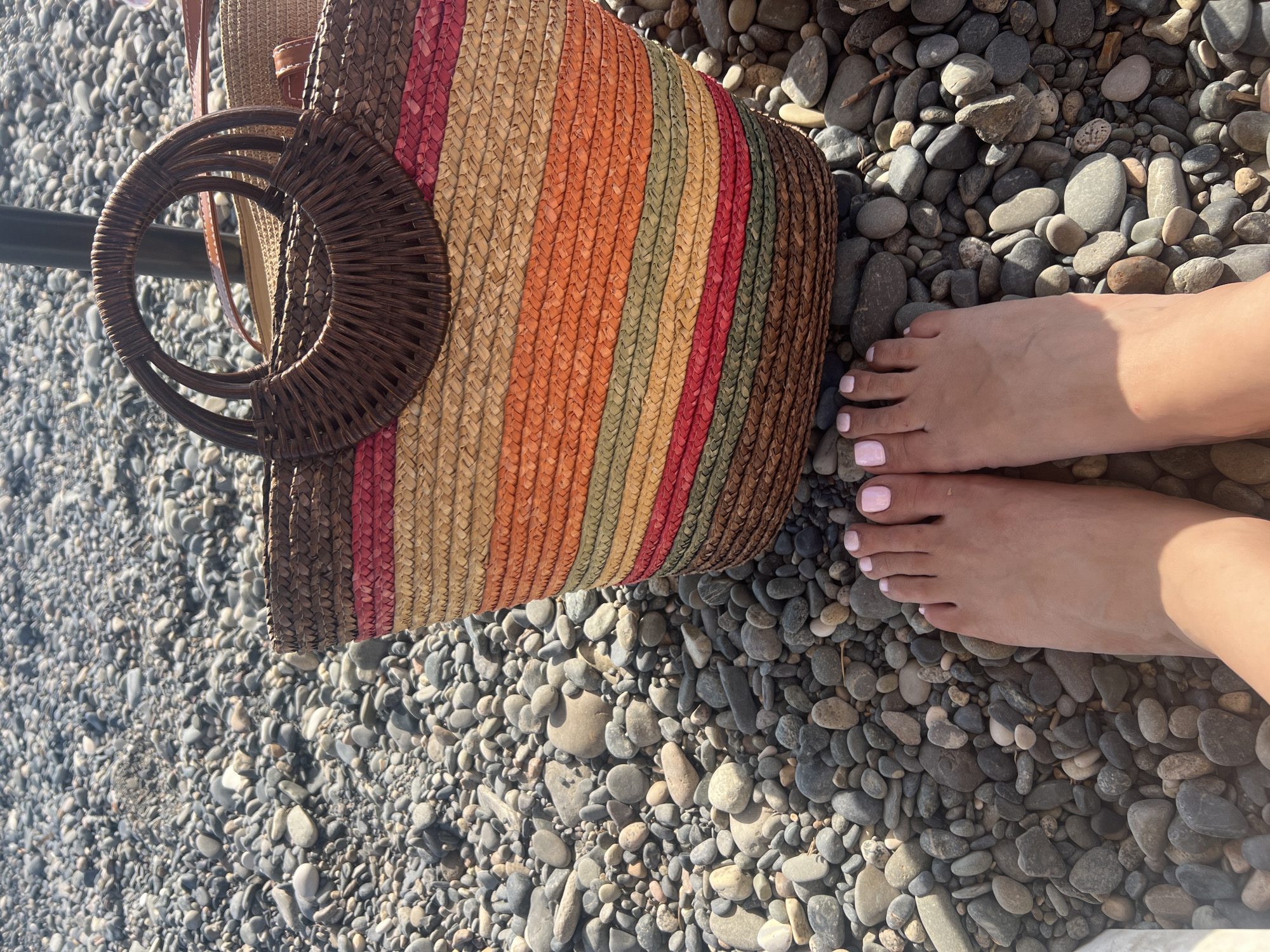 The image shows a top-down view of a straw beach bag resting on a pebble-strewn shore. The bag features colorful vertical stripes in shades of pink, orange, yellow, green, and brown, with dark wooden round handles. Next to the bag, a pair of bare feet with light pink nail polish are visible. The scene suggests a leisurely day at the beach, possibly in Sokhumi, Abkhazia, given the location context. The lighting indicates it is daytime, likely sunny, given the shadows. The pebbles on the shore are various sizes and shades of gray, brown, and white, typical of a coastal environment. There are no visible text elements in the image. The overall impression is one of relaxation and summer vacation.