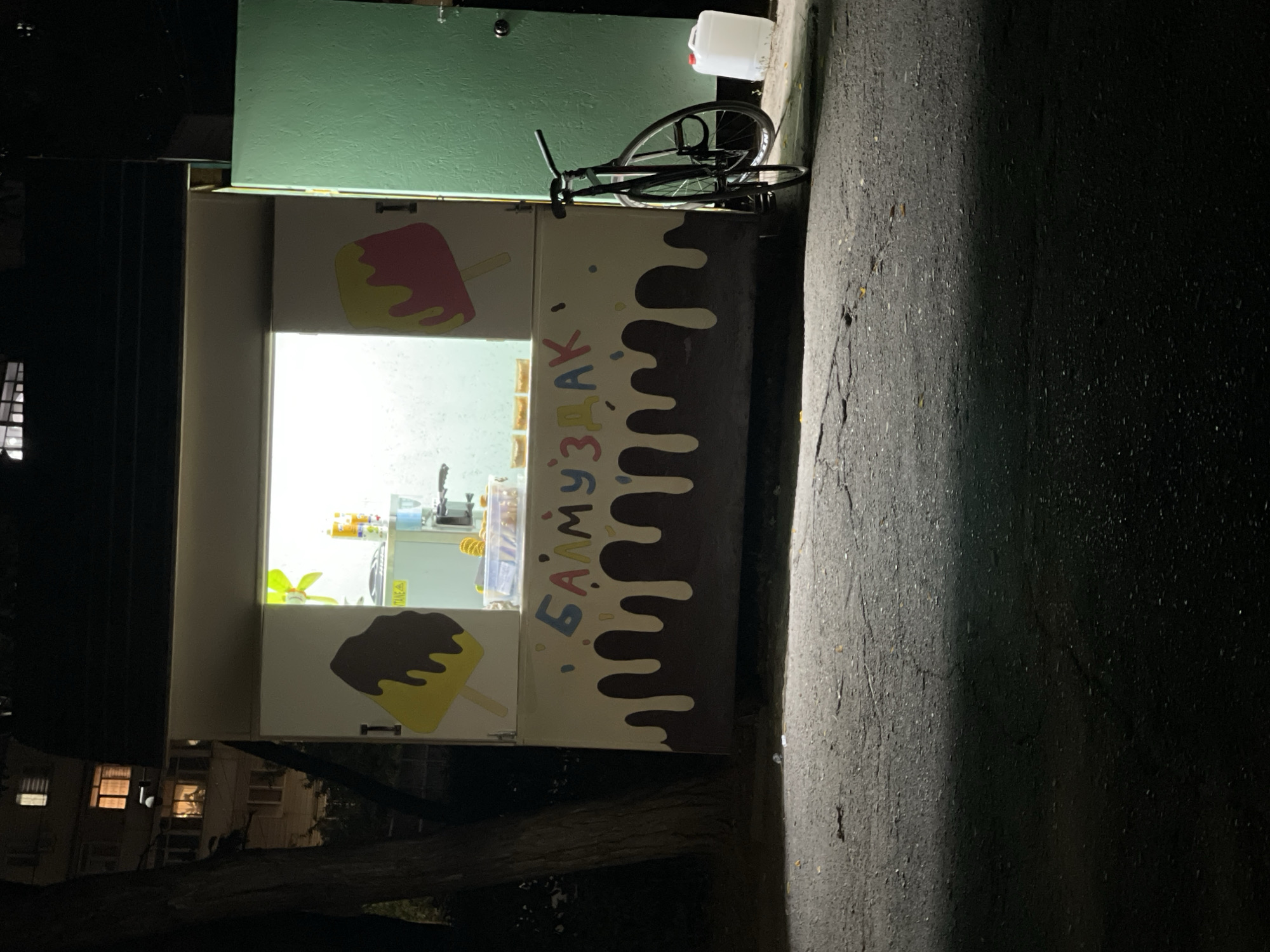 In Bishkek, Kyrgyzstan, at night, a small kiosk is illuminated with a bright interior light, displaying cartoon images of popsicles on its exterior. The kiosk is decorated with dripping chocolate-like patterns and colorful text that reads "БАЛМУЗДАК" (which translates to "ice cream" in Kyrgyz). A bicycle is parked leaning against the right side of the kiosk, near a closed green door. To the right of the bicycle, a white plastic container is visible on the ground. The kiosk appears to be situated on an asphalted area, possibly a sidewalk or a small parking lot, with a building visible in the background. The scene suggests a quiet, urban setting after business hours.