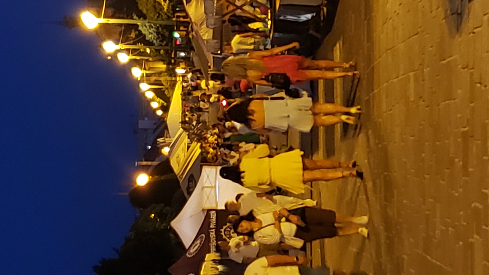 The image depicts an evening scene on a street in Prešov, Slovakia, likely at a festival or outdoor market. The sky is dark blue, indicating dusk or nighttime. Several streetlights with yellow bulbs line the right side of the image, illuminating a cobblestone pathway. On the left, white tents are set up, one displaying text that includes "PREŠOVSKÁ PIVOVÁREŇ," identifying it as related to a brewery in Prešov.
In the foreground, four women are walking away from the camera down the path. The woman on the far left wears a white top and dark shorts. Next to her, a woman in a bright yellow dress walks with her back to the camera. To her right, a woman in a light blue romper is also walking away. The woman on the far right wears a pink dress and carries a black bag over her shoulder.
In the background, a crowd of people are visible walking and gathered around stalls under more tents and lights. Trees are present on the left side of the image, and buildings are visible further in the distance. The overall atmosphere appears to be lively and social.