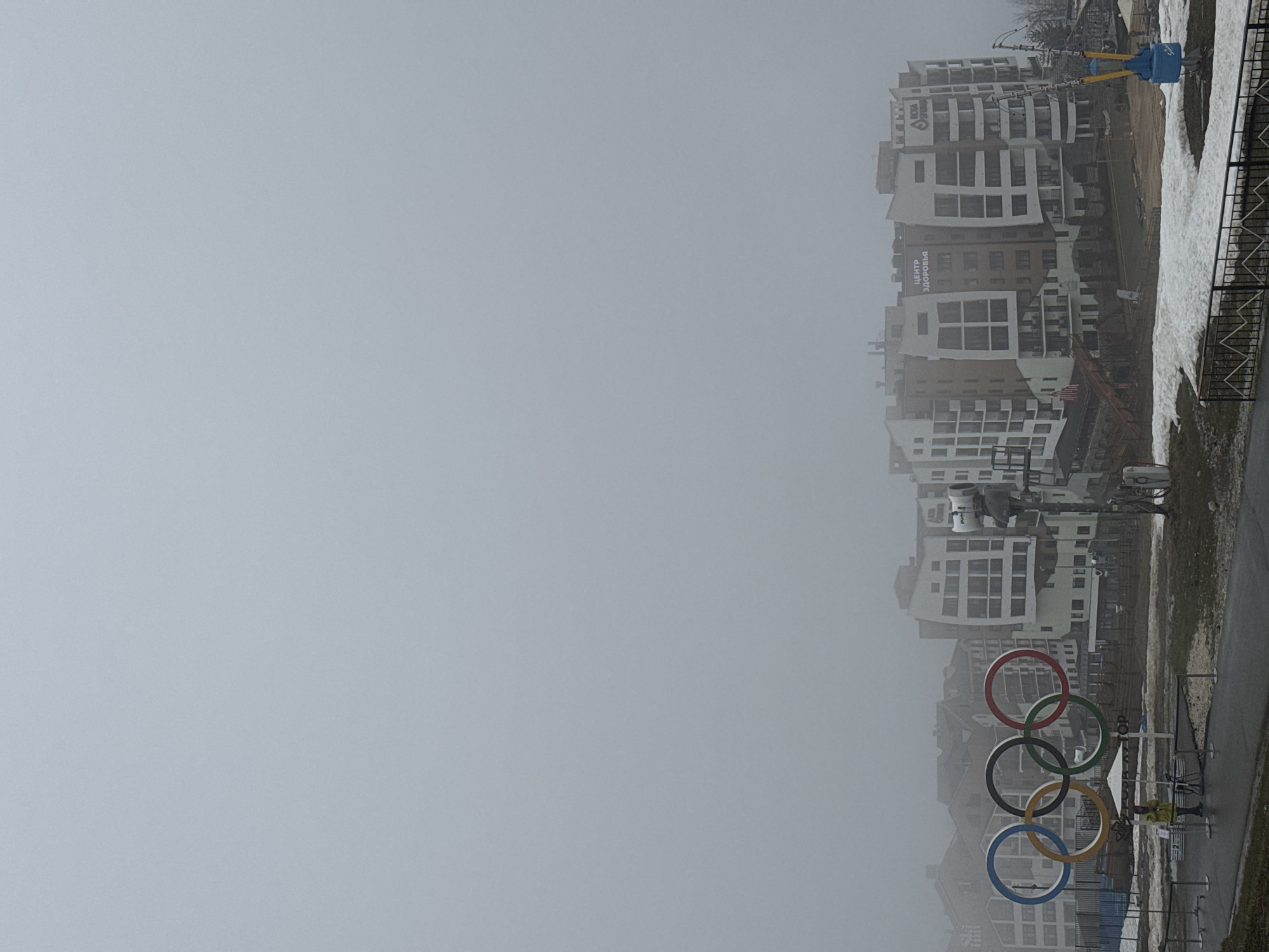 The image shows a hazy view of the Olympic rings in Сочи, Russia, with buildings visible in the background. The rings are in the standard Olympic colors, arranged in the correct order. The buildings in the background are tall and modern, with a mix of white and brown facades. The overall environment appears overcast and potentially snowy.