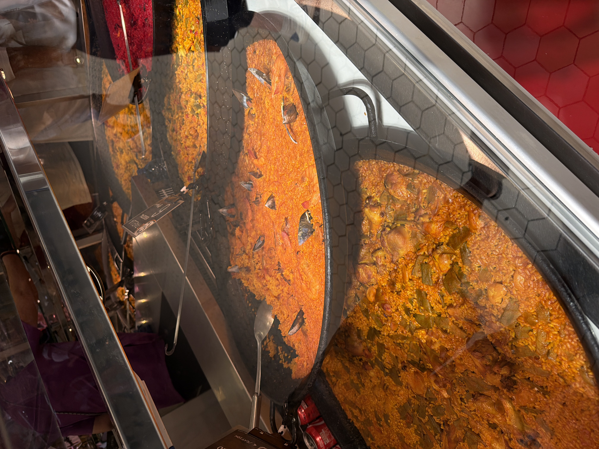 The image depicts an indoor food display, characteristic of a market or food hall, specifically featuring paella, a specialty of Valencia, Spain. The scene is dominated by multiple large, round paella pans filled with various preparations of the dish.
In the prominent foreground and mid-ground, a large, black, cast-iron paella pan is filled with a vibrant orange-red paella, likely a seafood variant, containing numerous cooked mussels (or similar bivalves) in their shells and possibly shrimp, distributed across the saffron-infused rice. A large, reflective silver serving spoon rests within this paella on the left side. Partially visible directly below this pan is another large, black paella pan containing an orange-brown paella with distinct pieces of chicken and elongated green bean segments (judía verde) mixed throughout the rice.
Further in the background, visible through the reflective glass display, are additional paella pans; one shows a yellow-orange paella, and another contains a bright red, possibly rice-based, dish. The food is showcased within a clear glass display case featuring silver metal framing, which reflects overhead lighting and structural elements of the environment.
Reflections on the glass also reveal two individuals: one on the far left wearing a purple top, and another on the right in a white garment, likely a uniform or apron. Two red Coca-Cola cans are visible on the left side, adjacent to the prominent paella pan. A black sign with partial white text, reading "PAELLA", is attached to the left side of the display structure. The floor on the far right of the image features a glossy red hexagonal tile pattern, while a similar darker hexagonal pattern is visible directly beneath the display case. The overall environment suggests a bustling commercial food service setting in the city of Valencia, Spain.